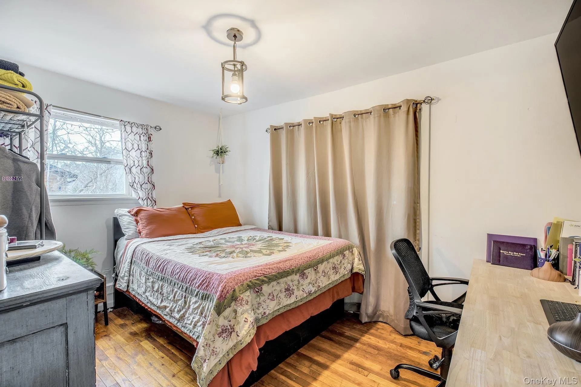 Bedroom featuring wood-finish flooring, a window with patterned drapes, a ceiling-mounted light fixture, and light-toned walls Bedroom featuring wood-finish flooring, a window with patterned drapes, a ceiling-mounted light fixture, and light-toned walls