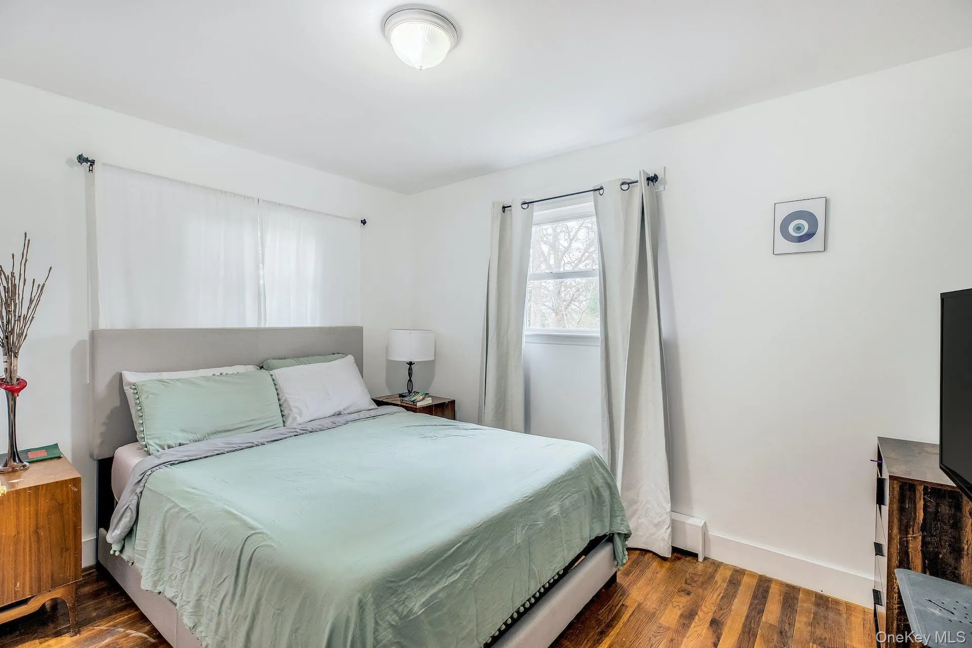 Bedroom featuring wood-finish flooring, white walls, and a recessed window with neutral drapes Bedroom featuring wood-finish flooring, white walls, and a recessed window with neutral drapes