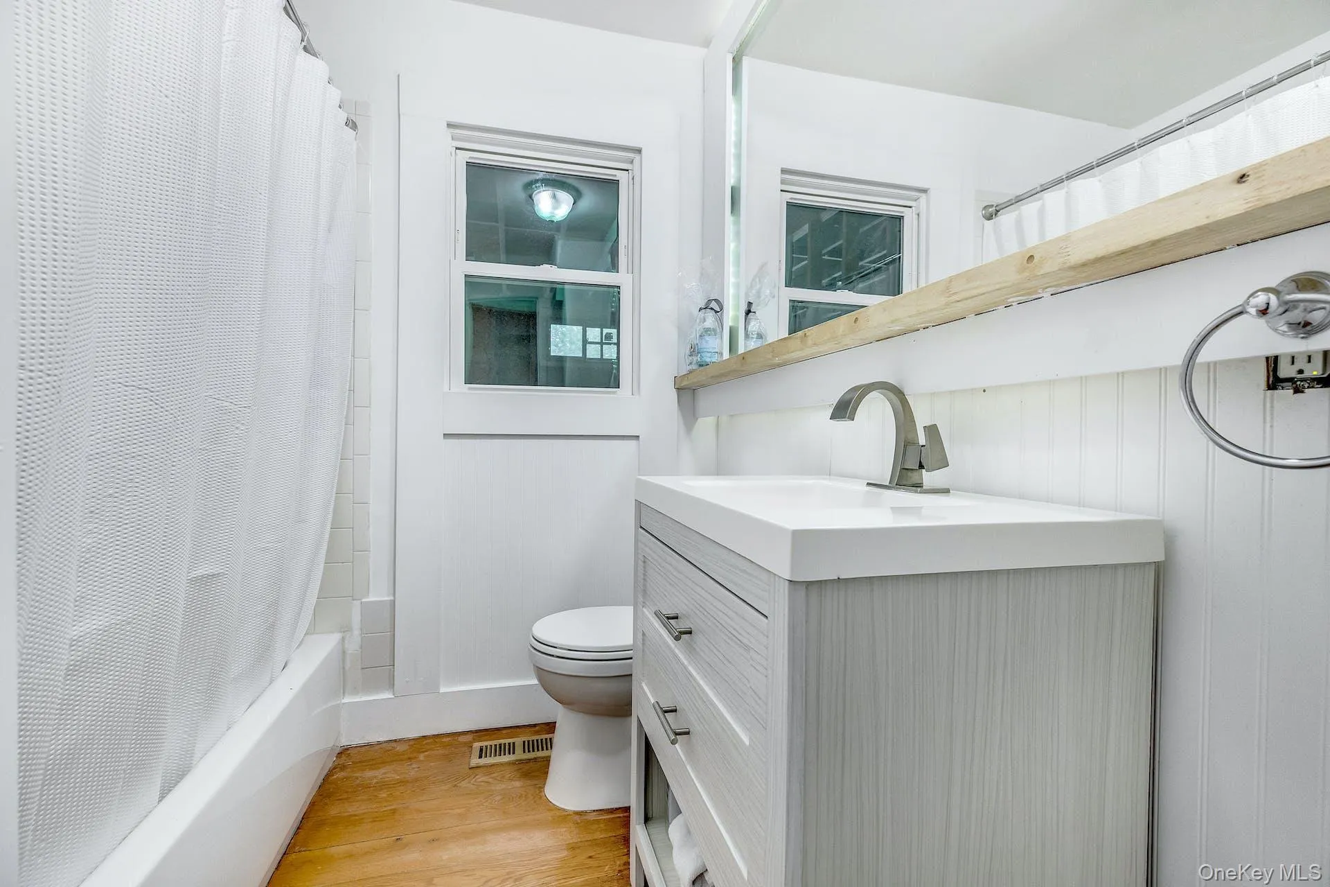 Bathroom featuring a single vanity with a white countertop and brushed nickel faucet, a window, a white bathtub with subway tile surround, white beadboard wainscoting, and wood-finish flooring Bathroom featuring a single vanity with a white countertop and brushed nickel faucet, a window, a white bathtub with subway tile surround, white beadboard wainscoting, and wood-finish flooring