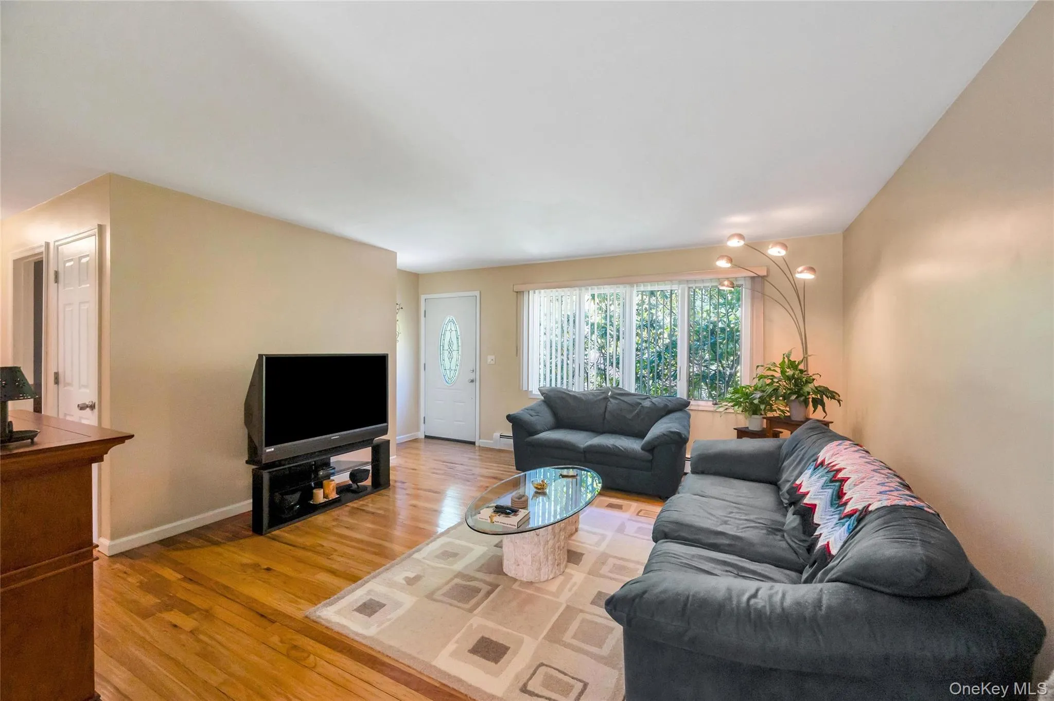 Living room featuring wood-finish flooring, neutral wall tones, a large front-facing window, and a white entry door with decorative glass inlay Living room featuring wood-finish flooring, neutral wall tones, a large front-facing window, and a white entry door with decorative glass inlay