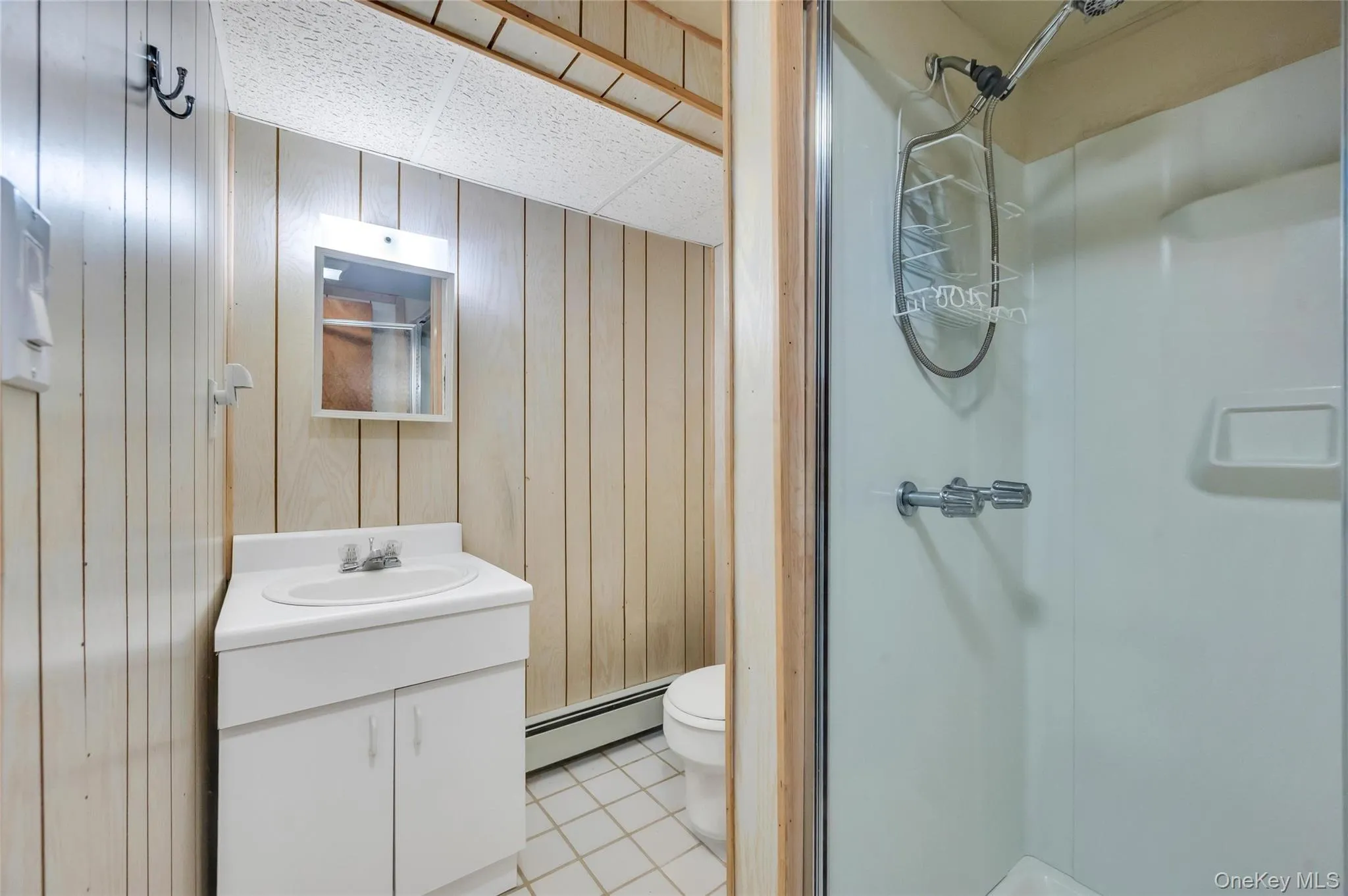 Bathroom featuring a white vanity with integrated sink, paneling walls, a mirrored medicine cabinet, and a shower stall with a clear glass enclosure Bathroom featuring a white vanity with integrated sink, paneling walls, a mirrored medicine cabinet, and a shower stall with a clear glass enclosure