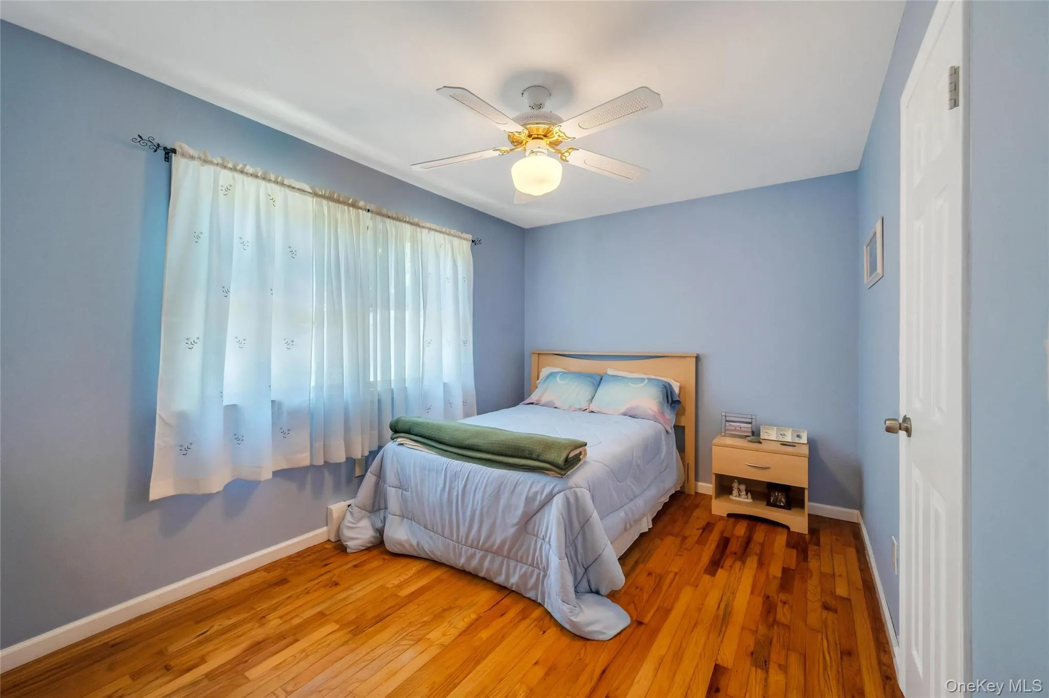 Hardwood flooring and light blue walls define this room, featuring a window with sheer curtains, a white ceiling fan with integrated lighting, and a paneled white door Hardwood flooring and light blue walls define this room, featuring a window with sheer curtains, a white ceiling fan with integrated lighting, and a paneled white door