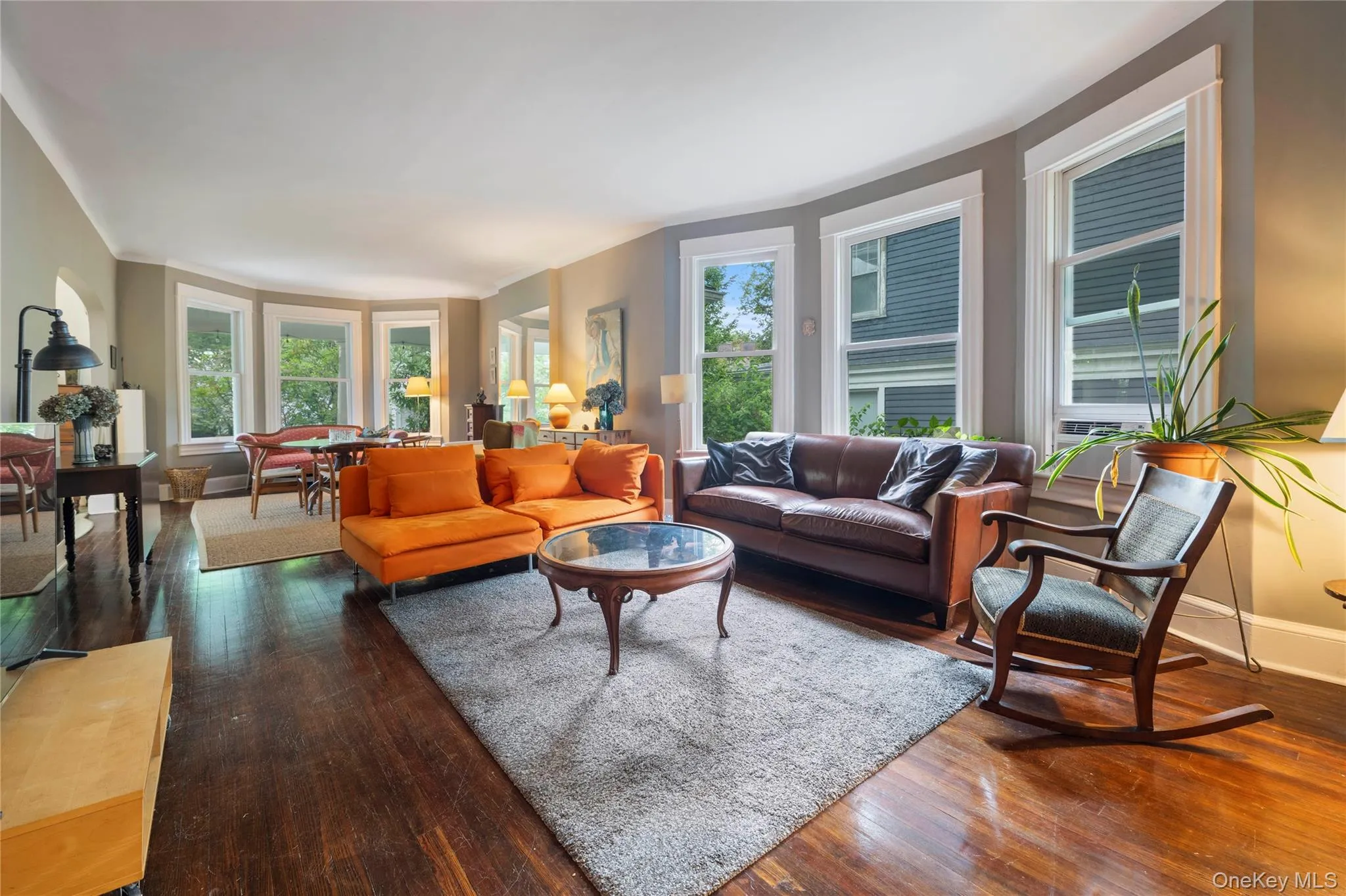 Spacious living area featuring polished wood-finish flooring, a curved wall of multiple windows with white trim, and a wide entryway Spacious living area featuring polished wood-finish flooring, a curved wall of multiple windows with white trim, and a wide entryway