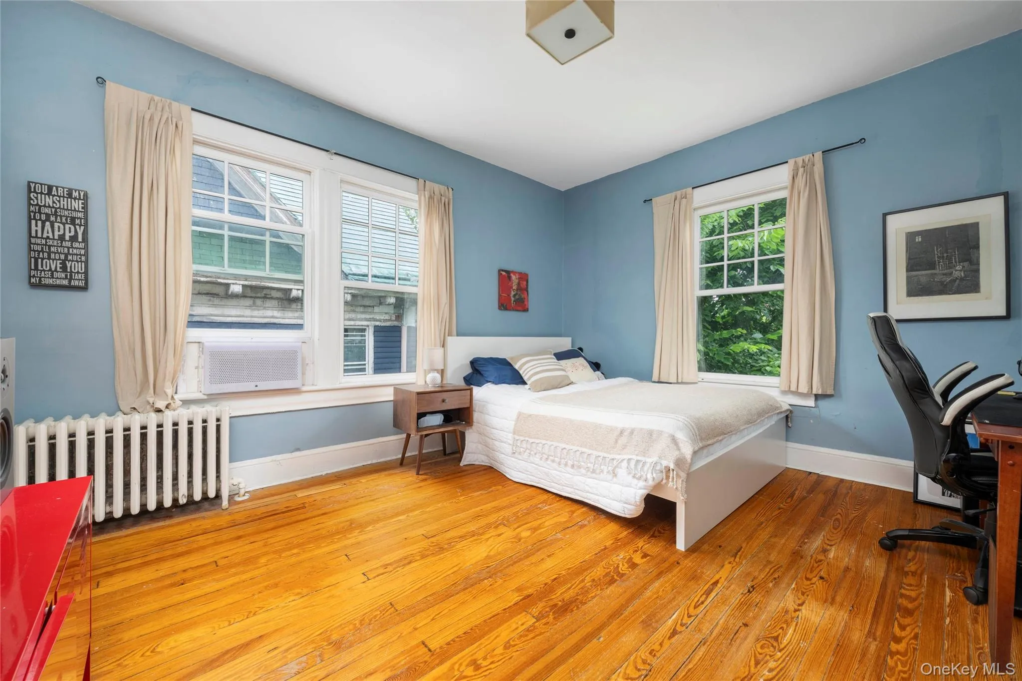 Room featuring wood-finish flooring, two large windows with white trim, and a ceiling-mounted light fixture Room featuring wood-finish flooring, two large windows with white trim, and a ceiling-mounted light fixture