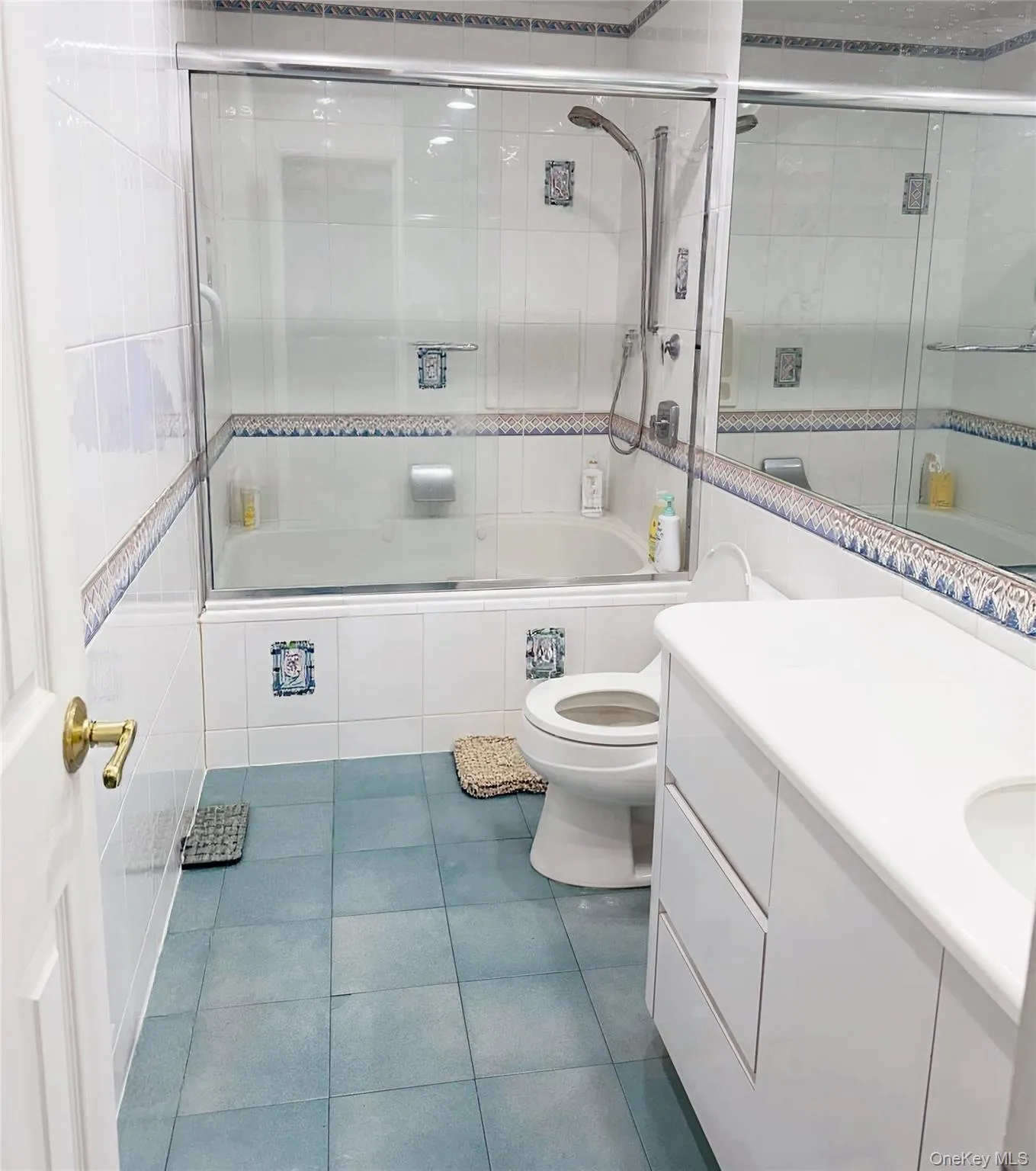 Bathroom featuring a white tile shower surround with decorative accents, a glass sliding shower door over a bathtub, a white vanity with integrated sink, and light blue floor tiles Bathroom featuring a white tile shower surround with decorative accents, a glass sliding shower door over a bathtub, a white vanity with integrated sink, and light blue floor tiles