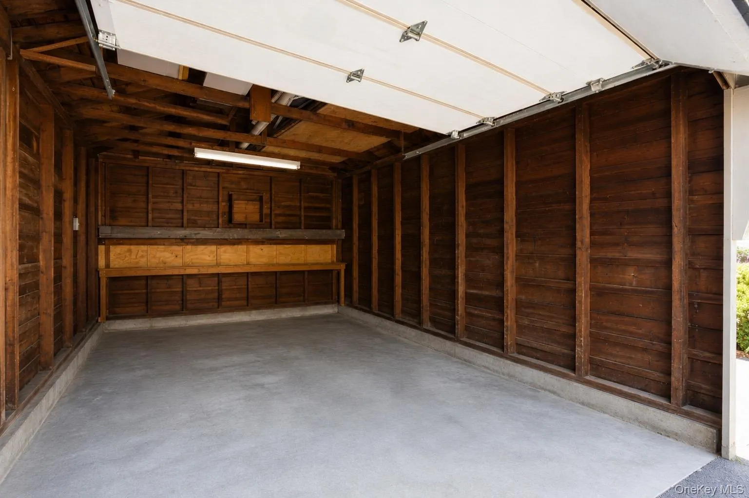 Spacious garage featuring a smooth concrete floor, natural wood-paneled walls, exposed wood beam ceiling, built-in shelving, and overhead fluorescent lighting Spacious garage featuring a smooth concrete floor, natural wood-paneled walls, exposed wood beam ceiling, built-in shelving, and overhead fluorescent lighting