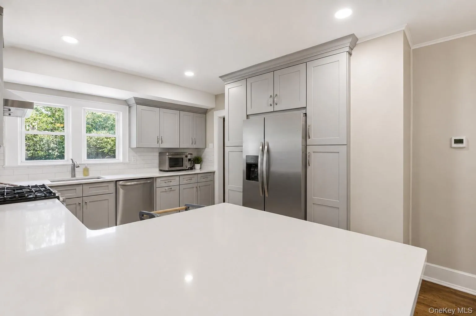 Modern kitchen featuring light gray cabinetry, stainless steel appliances, white subway tile backsplash, recessed lighting, and a spacious white countertop with a gas cooktop Modern kitchen featuring light gray cabinetry, stainless steel appliances, white subway tile backsplash, recessed lighting, and a spacious white countertop with a gas cooktop