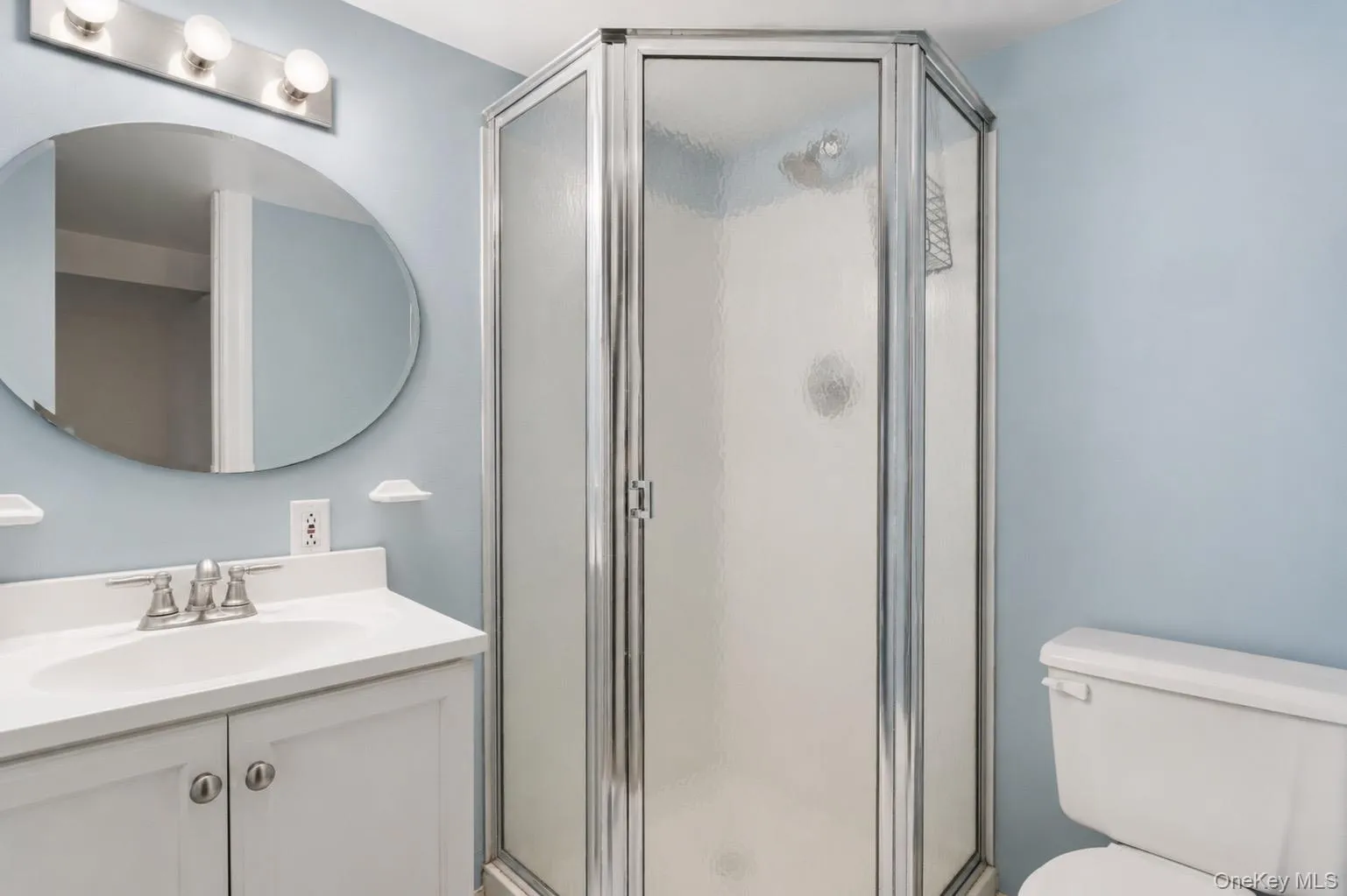 Hexagonal glass shower enclosure with chrome framing, a white vanity with an integrated sink, brushed nickel faucet, and an oval wall mirror with a three-light fixture Hexagonal glass shower enclosure with chrome framing, a white vanity with an integrated sink, brushed nickel faucet, and an oval wall mirror with a three-light fixture
