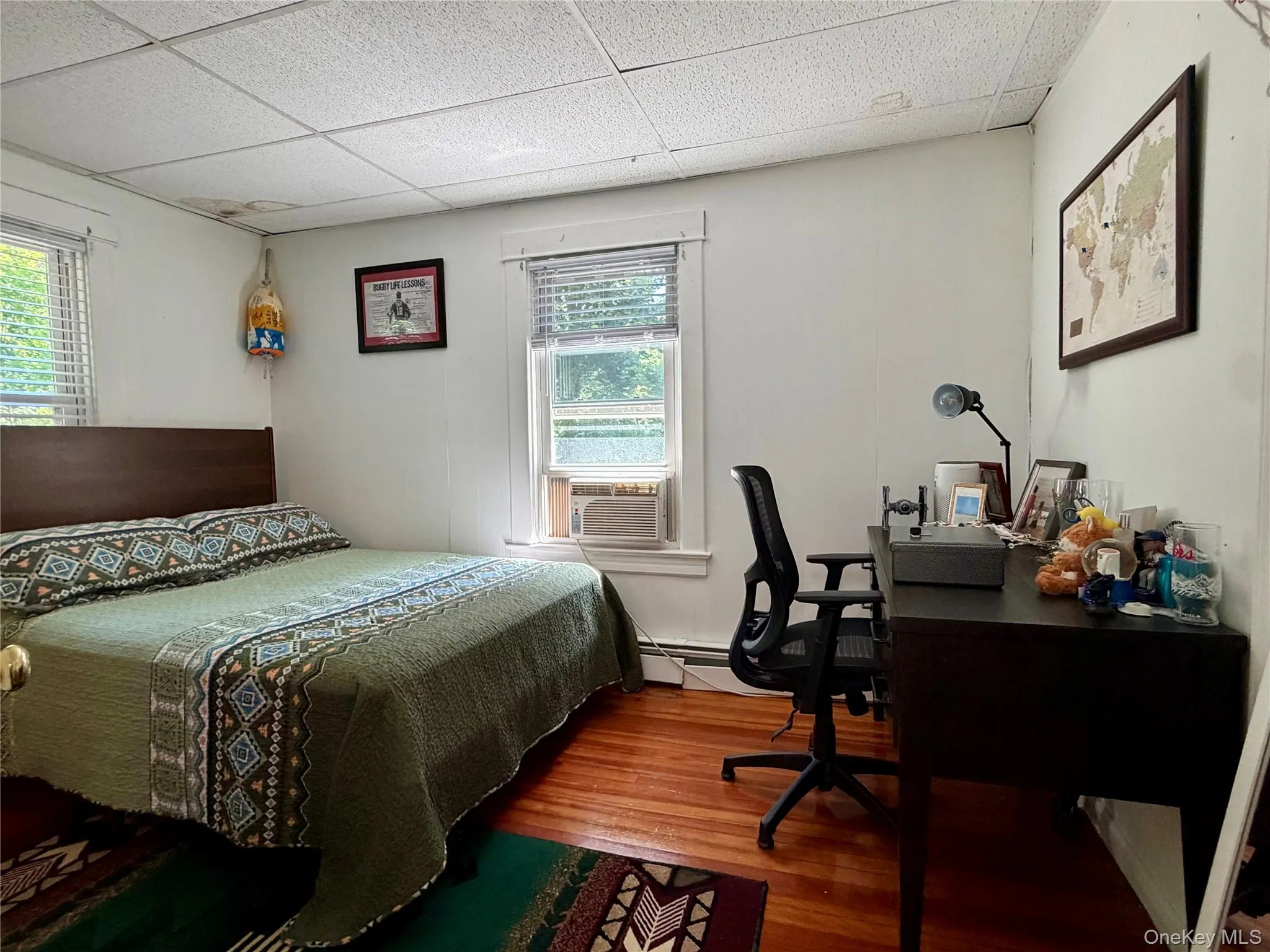 Room featuring wood-finish flooring, two windows with blinds, a paneled ceiling, and white wall finishes Room featuring wood-finish flooring, two windows with blinds, a paneled ceiling, and white wall finishes