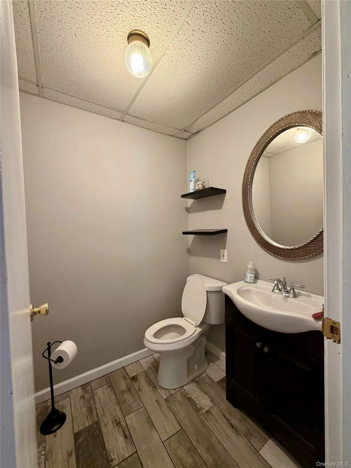Bathroom featuring wood-finish flooring, a contemporary vanity with an integrated sink, a toilet, a decorative round mirror, and a drop ceiling with recessed lighting Bathroom featuring wood-finish flooring, a contemporary vanity with an integrated sink, a toilet, a decorative round mirror, and a drop ceiling with recessed lighting