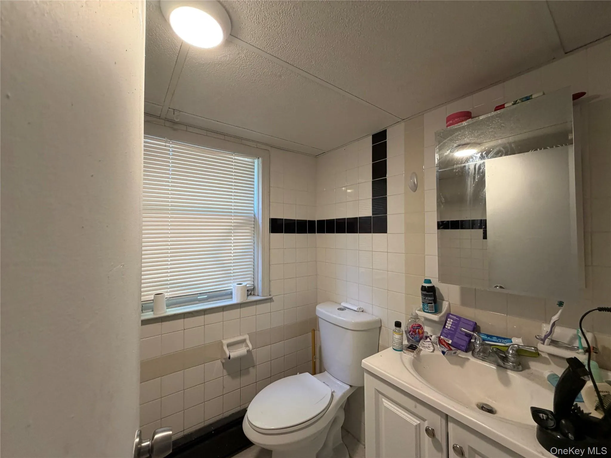 Bathroom featuring white wall tiling with black accent bands, a window with horizontal blinds, a white vanity with cabinet storage, and a wall-mounted mirror Bathroom featuring white wall tiling with black accent bands, a window with horizontal blinds, a white vanity with cabinet storage, and a wall-mounted mirror