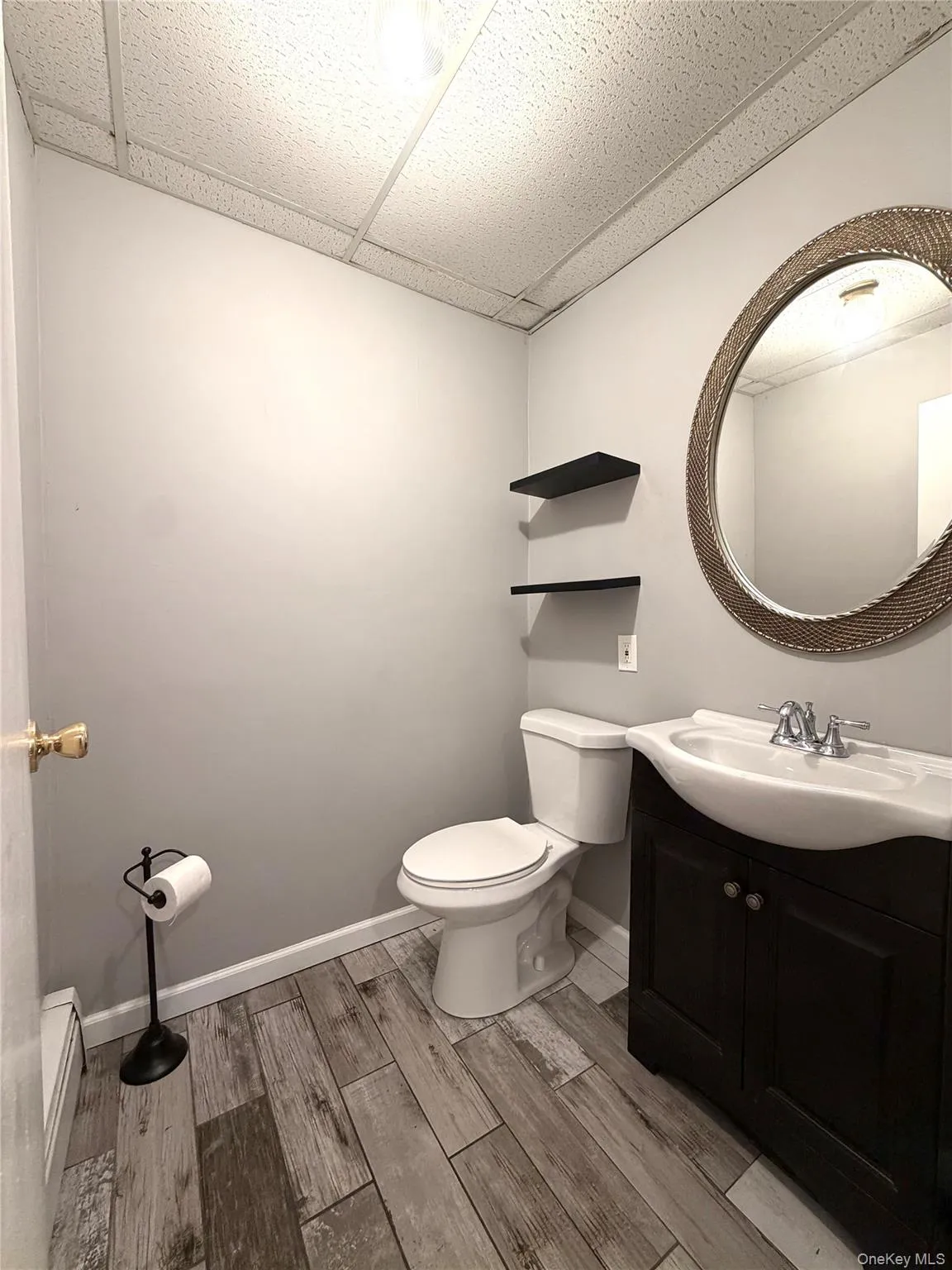 Wood-finish flooring, white toilet, dark wood vanity, white sink, and round wall-mounted mirror Wood-finish flooring, white toilet, dark wood vanity, white sink, and round wall-mounted mirror