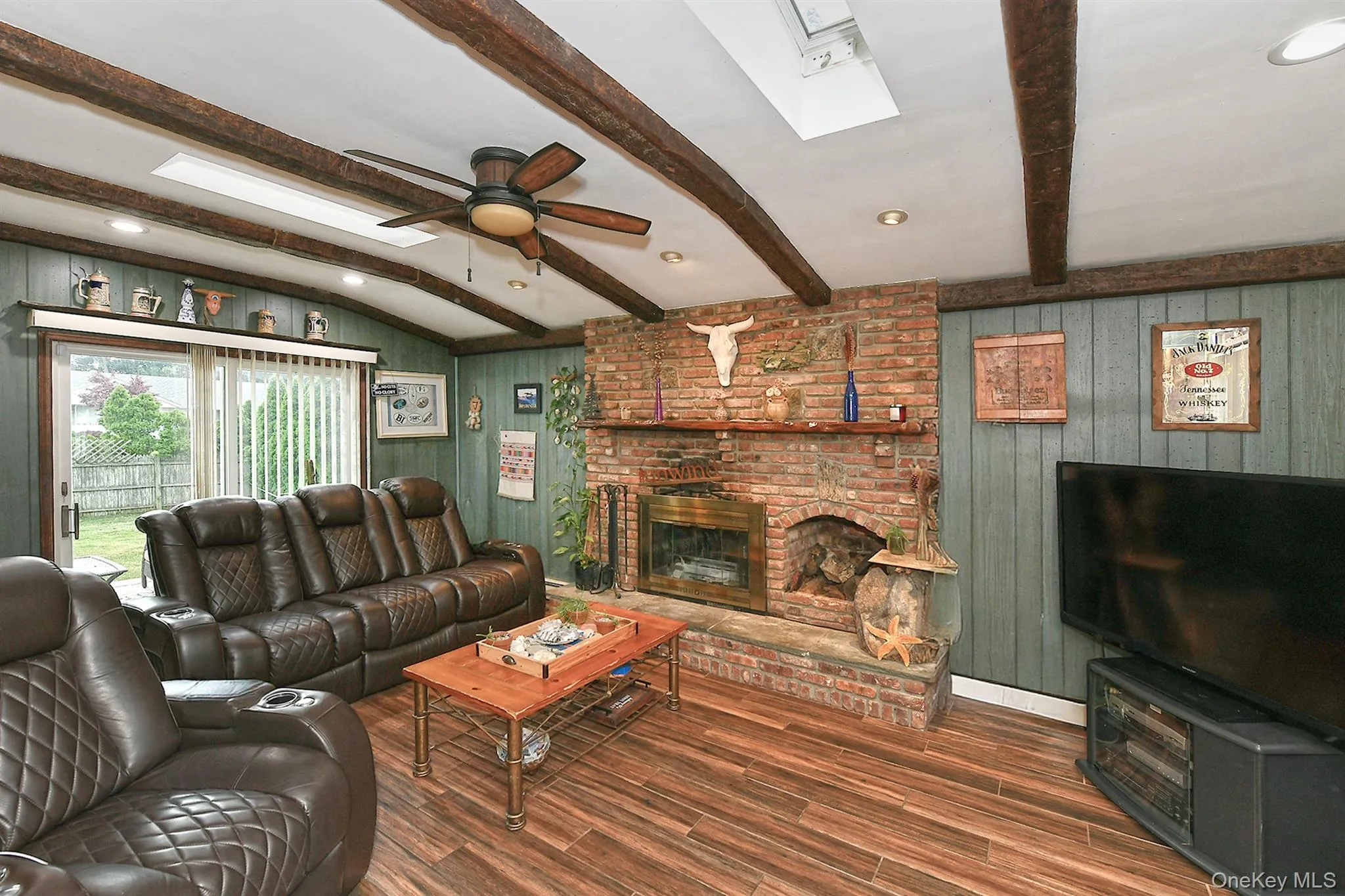 Living area featuring a brick fireplace, wood-finish flooring, exposed ceiling beams, and a ceiling fan Living area featuring a brick fireplace, wood-finish flooring, exposed ceiling beams, and a ceiling fan