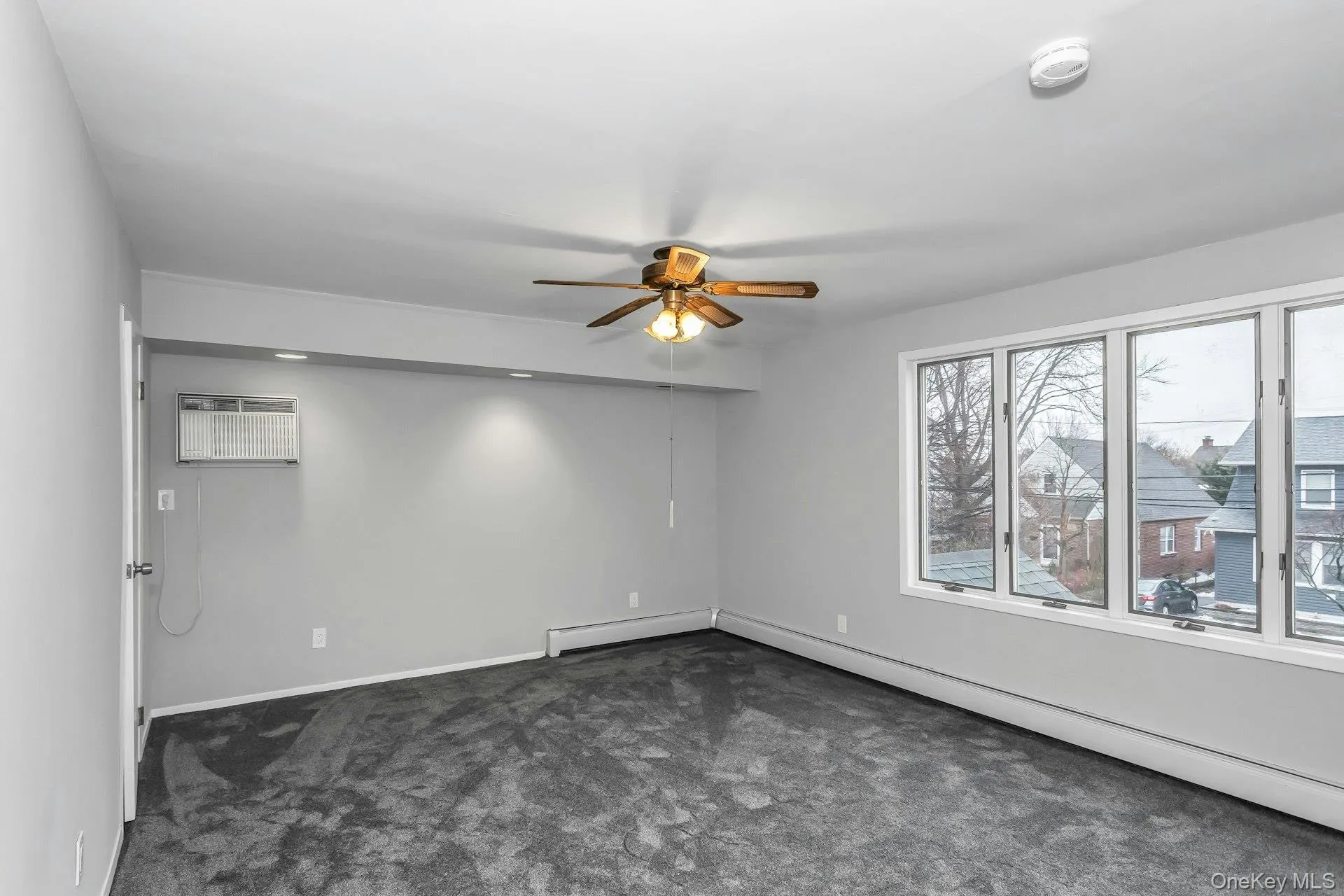 Spacious room featuring light gray walls, dark gray carpet, a multi-pane window, a ceiling fan with integrated lighting, and an air conditioning unit Spacious room featuring light gray walls, dark gray carpet, a multi-pane window, a ceiling fan with integrated lighting, and an air conditioning unit