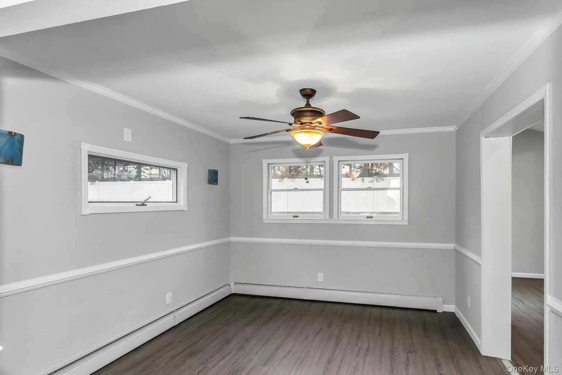 Spacious room featuring wood-finish flooring, light gray walls with white chair rail molding, and a bronze ceiling fan with integrated lighting Spacious room featuring wood-finish flooring, light gray walls with white chair rail molding, and a bronze ceiling fan with integrated lighting