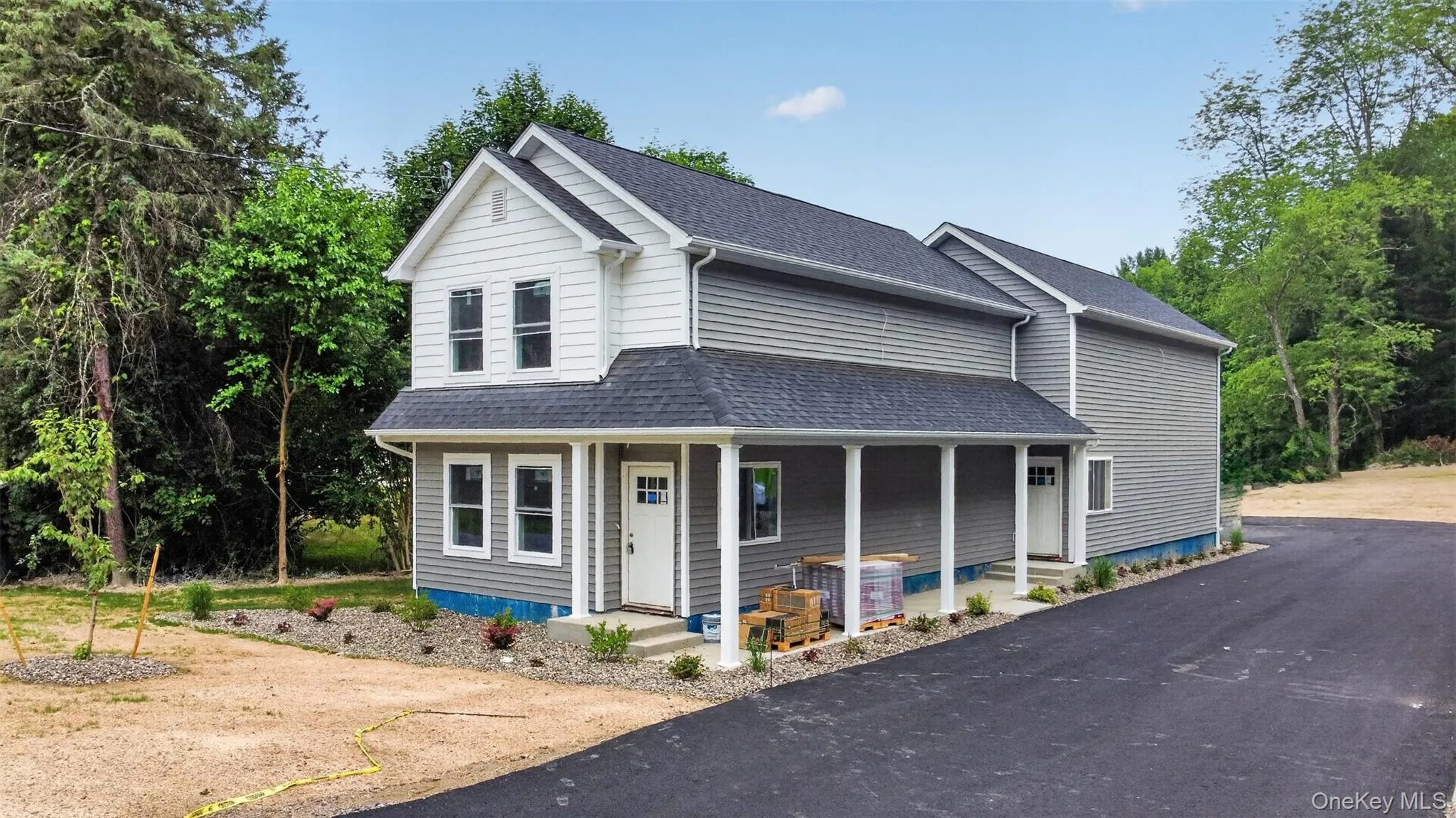 Newly constructed residence featuring a gray and white siding exterior, dark shingle roofing, a covered porch with white columns, a paved driveway, and nascent landscaping Newly constructed residence featuring a gray and white siding exterior, dark shingle roofing, a covered porch with white columns, a paved driveway, and nascent landscaping