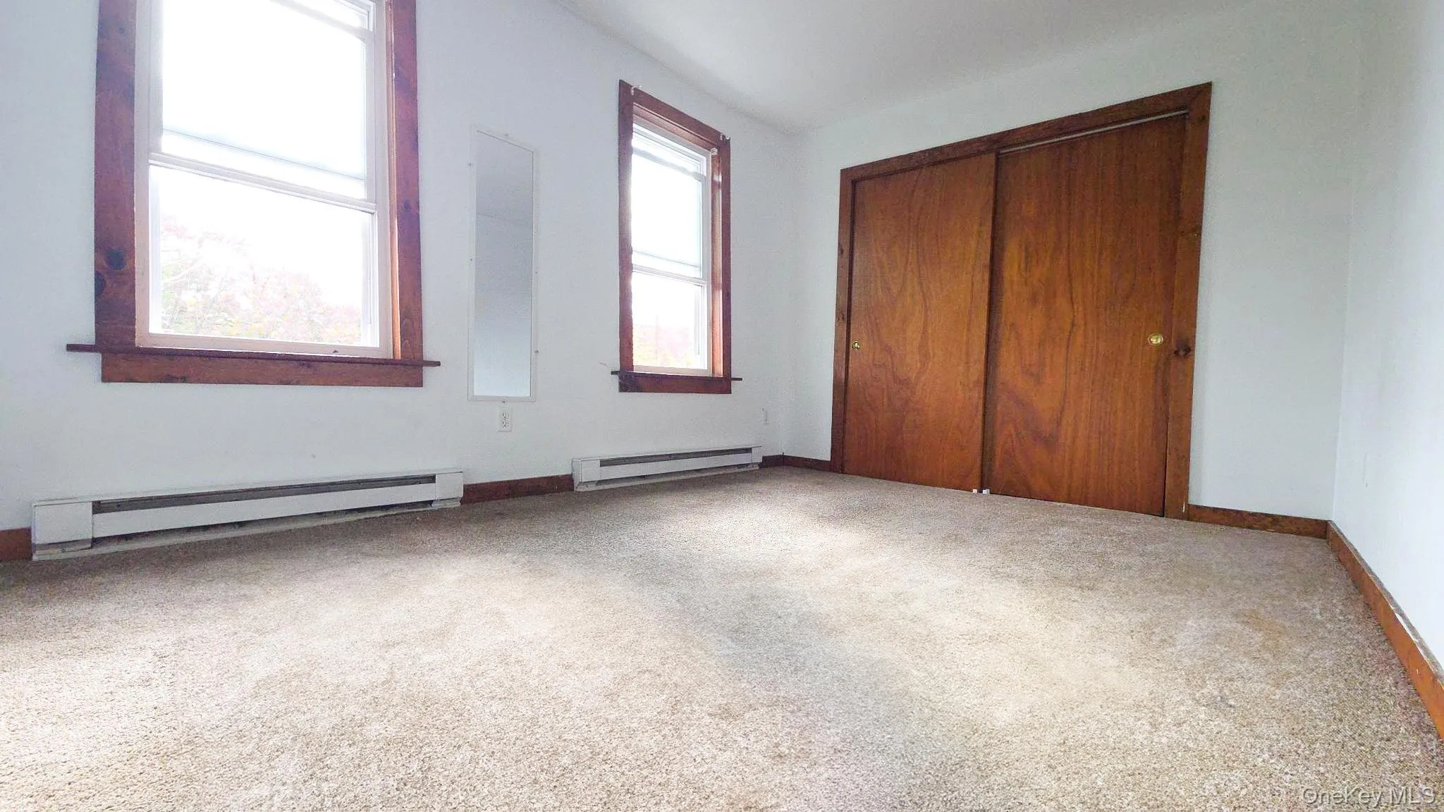 Carpeted room featuring two windows with wood trim, a built-in closet with wood sliding doors, and two baseboard heaters Carpeted room featuring two windows with wood trim, a built-in closet with wood sliding doors, and two baseboard heaters
