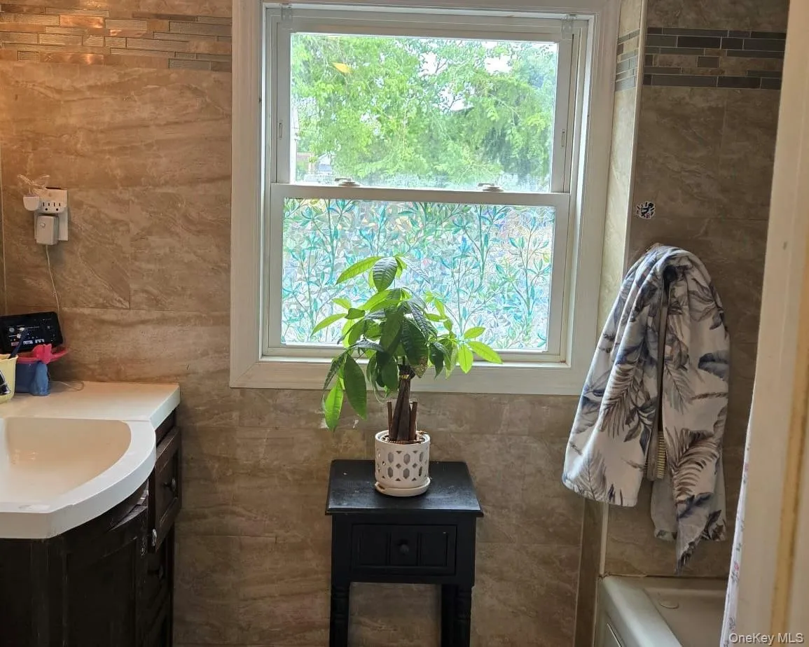 Bathroom featuring full-height stone-finish wall tile, a single-hung window with privacy film, a dark wood-finish vanity with a white countertop, and a built-in bathtub Bathroom featuring full-height stone-finish wall tile, a single-hung window with privacy film, a dark wood-finish vanity with a white countertop, and a built-in bathtub