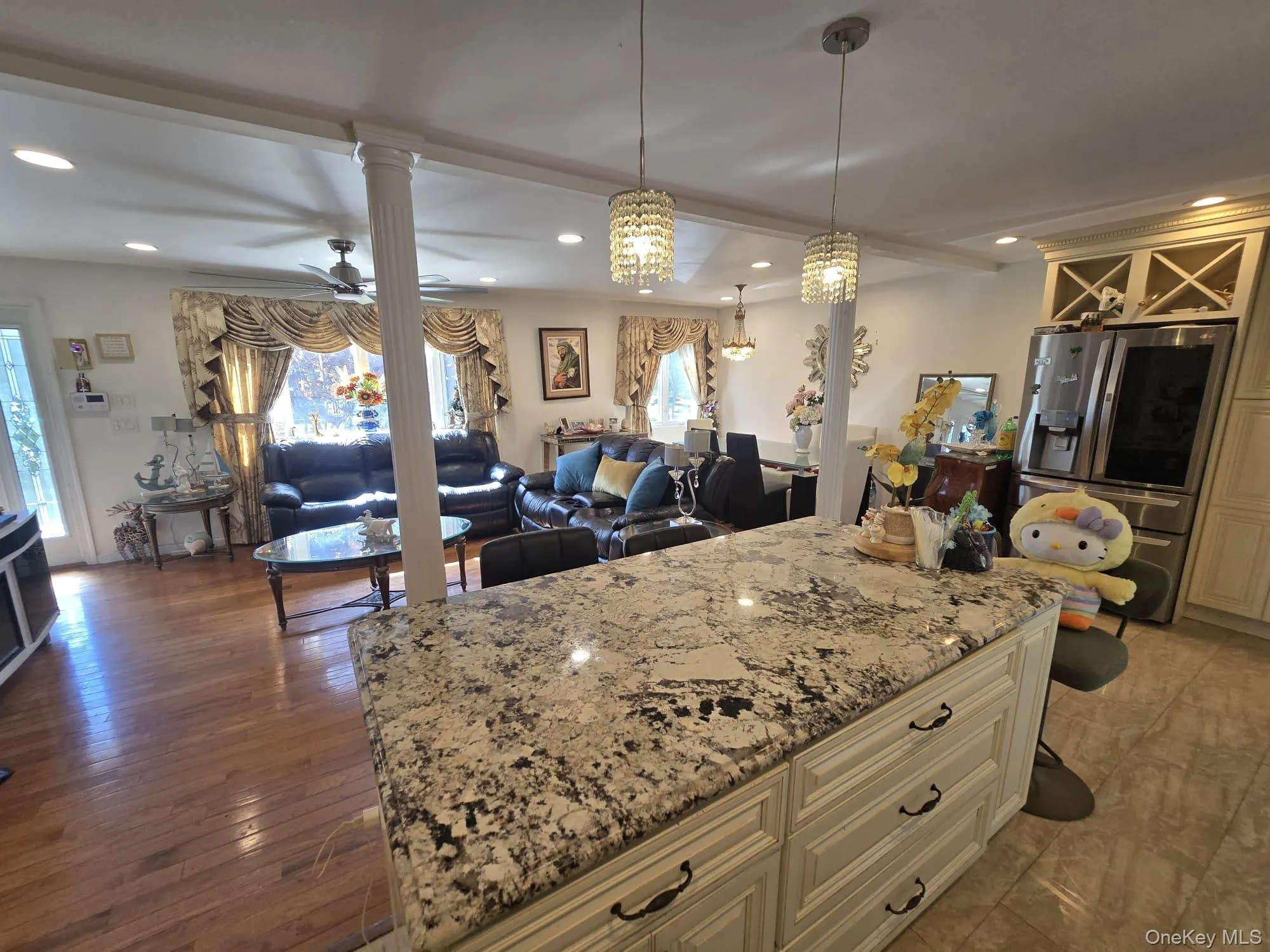 Spacious open-concept interior featuring a large kitchen island with a speckled countertop and cream-colored cabinetry, two crystal pendant lights, recessed ceiling lighting, and wood-finish flooring extending into the living area Spacious open-concept interior featuring a large kitchen island with a speckled countertop and cream-colored cabinetry, two crystal pendant lights, recessed ceiling lighting, and wood-finish flooring extending into the living area