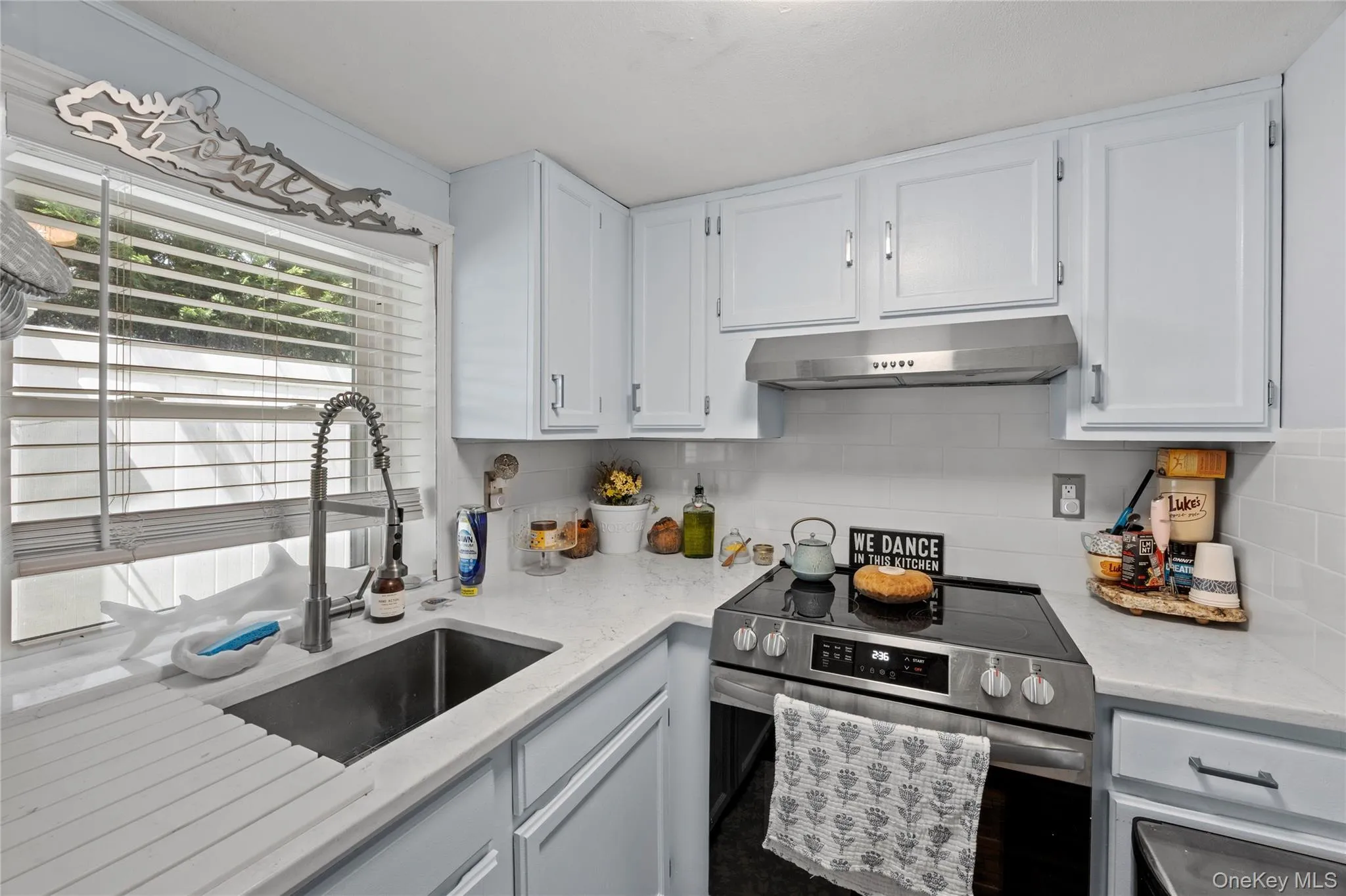 Modern kitchen featuring light blue cabinetry with brushed nickel hardware, a stainless steel range with overhead ventilation, a single basin stainless steel sink with a gooseneck faucet, light-toned countertops, and a white subway tile backsplash Modern kitchen featuring light blue cabinetry with brushed nickel hardware, a stainless steel range with overhead ventilation, a single basin stainless steel sink with a gooseneck faucet, light-toned countertops, and a white subway tile backsplash