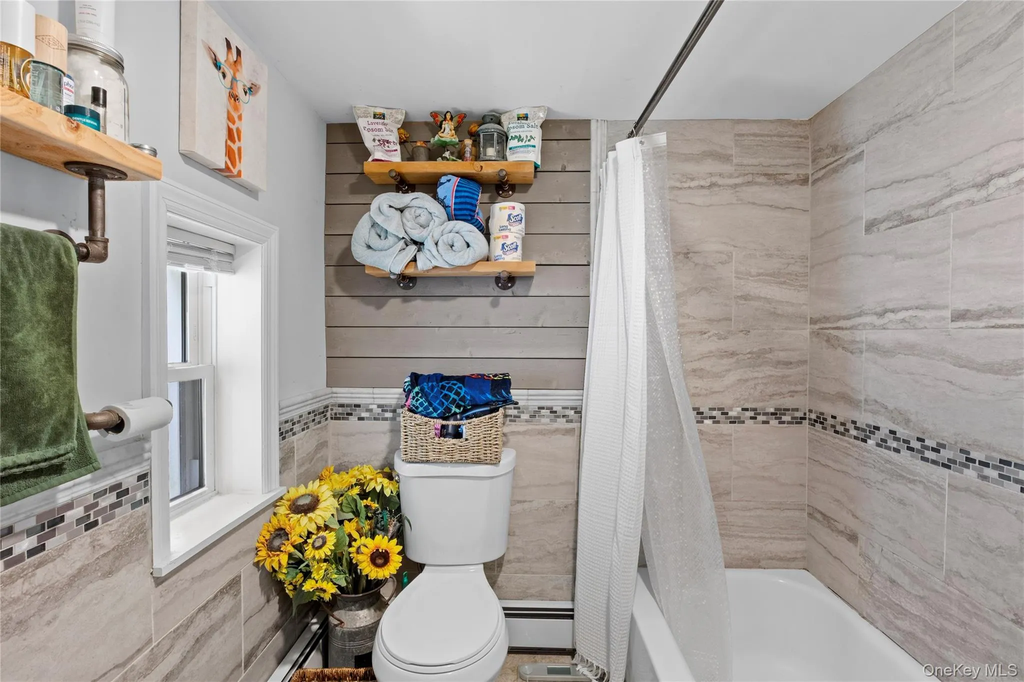 Bathroom featuring a white bathtub, light-toned tile shower surround with a decorative mosaic band, and a window with white trim Bathroom featuring a white bathtub, light-toned tile shower surround with a decorative mosaic band, and a window with white trim