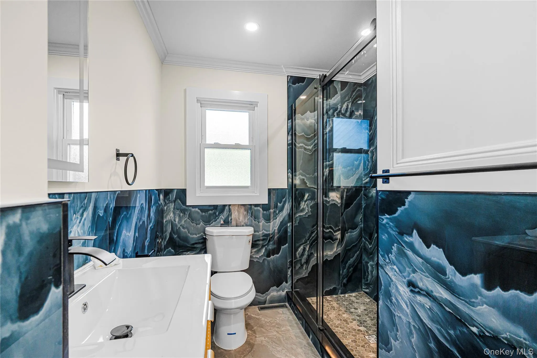 Bathroom featuring blue wave-patterned wall tiling, a glass-enclosed shower with a pebble-tiled floor, and a white vanity with a single basin and a matte black faucet Bathroom featuring blue wave-patterned wall tiling, a glass-enclosed shower with a pebble-tiled floor, and a white vanity with a single basin and a matte black faucet
