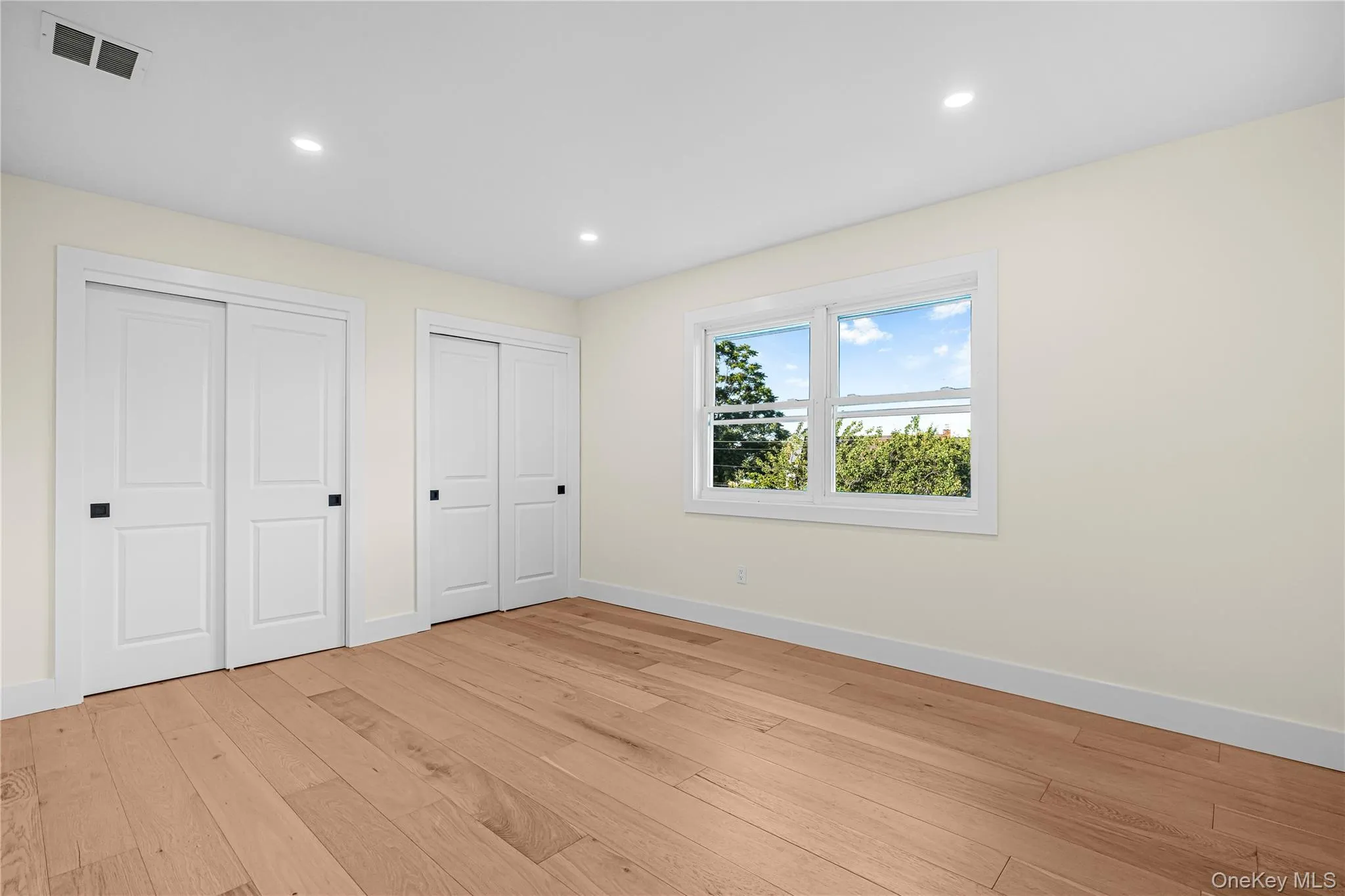 Spacious room featuring wood-finish flooring, two sets of bi-fold closet doors with black hardware, a large window, recessed lighting, and white baseboards Spacious room featuring wood-finish flooring, two sets of bi-fold closet doors with black hardware, a large window, recessed lighting, and white baseboards