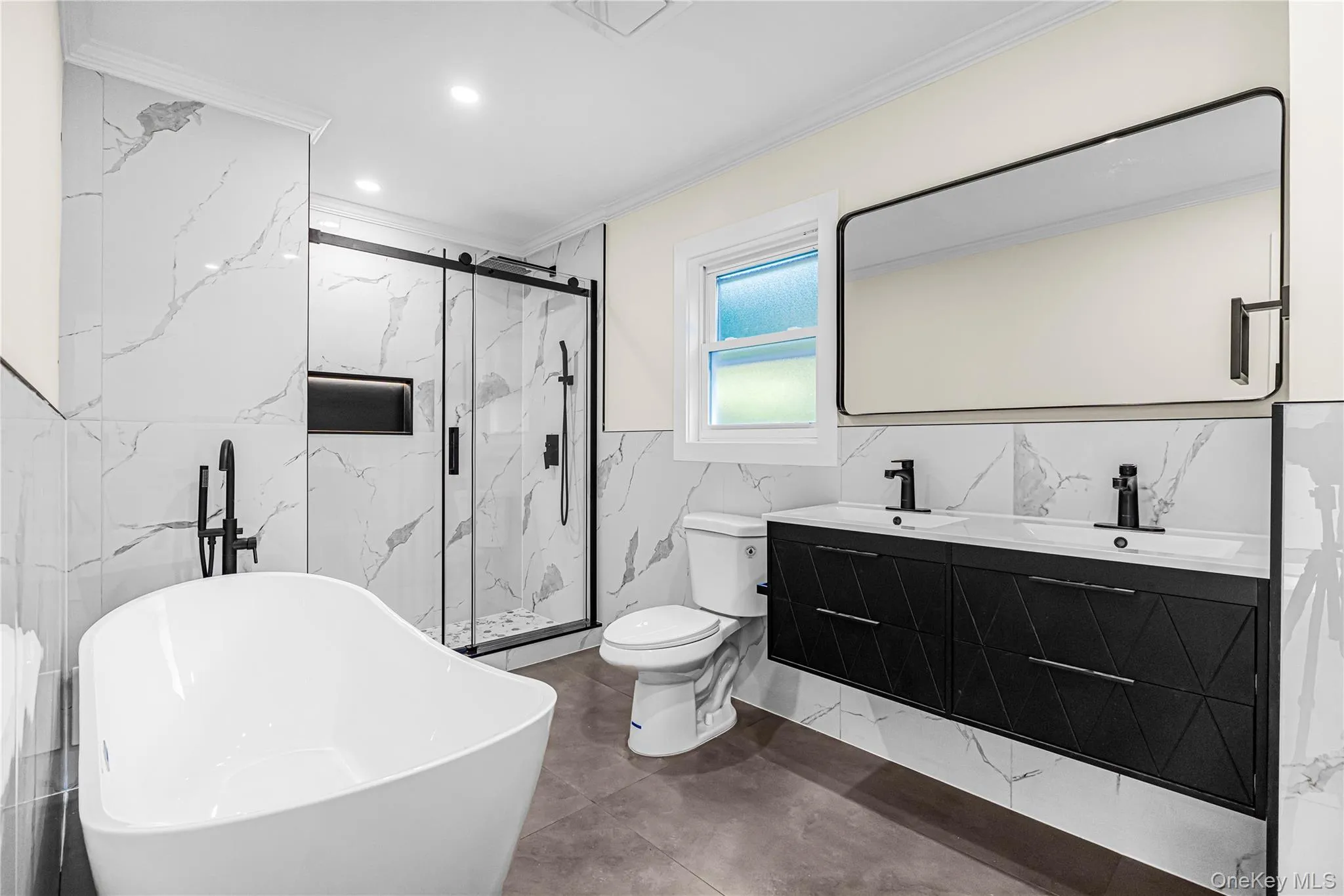 Contemporary bathroom featuring a freestanding tub with a matte black floor-mounted faucet, a double vanity with matte black fixtures, a walk-in shower with a glass enclosure, and white marble-style wall tiling Contemporary bathroom featuring a freestanding tub with a matte black floor-mounted faucet, a double vanity with matte black fixtures, a walk-in shower with a glass enclosure, and white marble-style wall tiling
