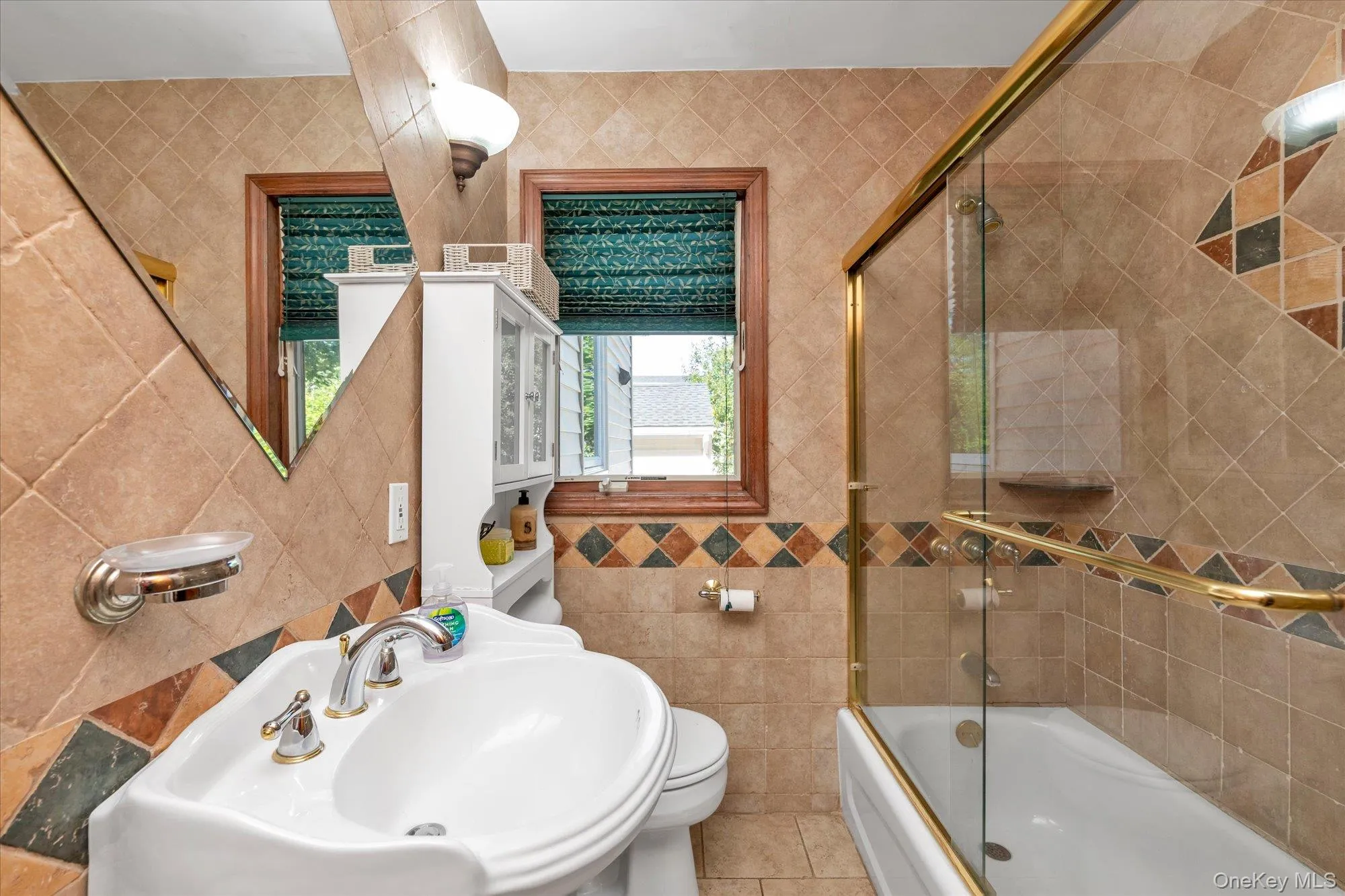 Bathroom featuring a pedestal sink with dual-handle faucet, full-height tiled walls, a built-in medicine cabinet, a window with wood trim, and a bathtub with a sliding glass enclosure Bathroom featuring a pedestal sink with dual-handle faucet, full-height tiled walls, a built-in medicine cabinet, a window with wood trim, and a bathtub with a sliding glass enclosure