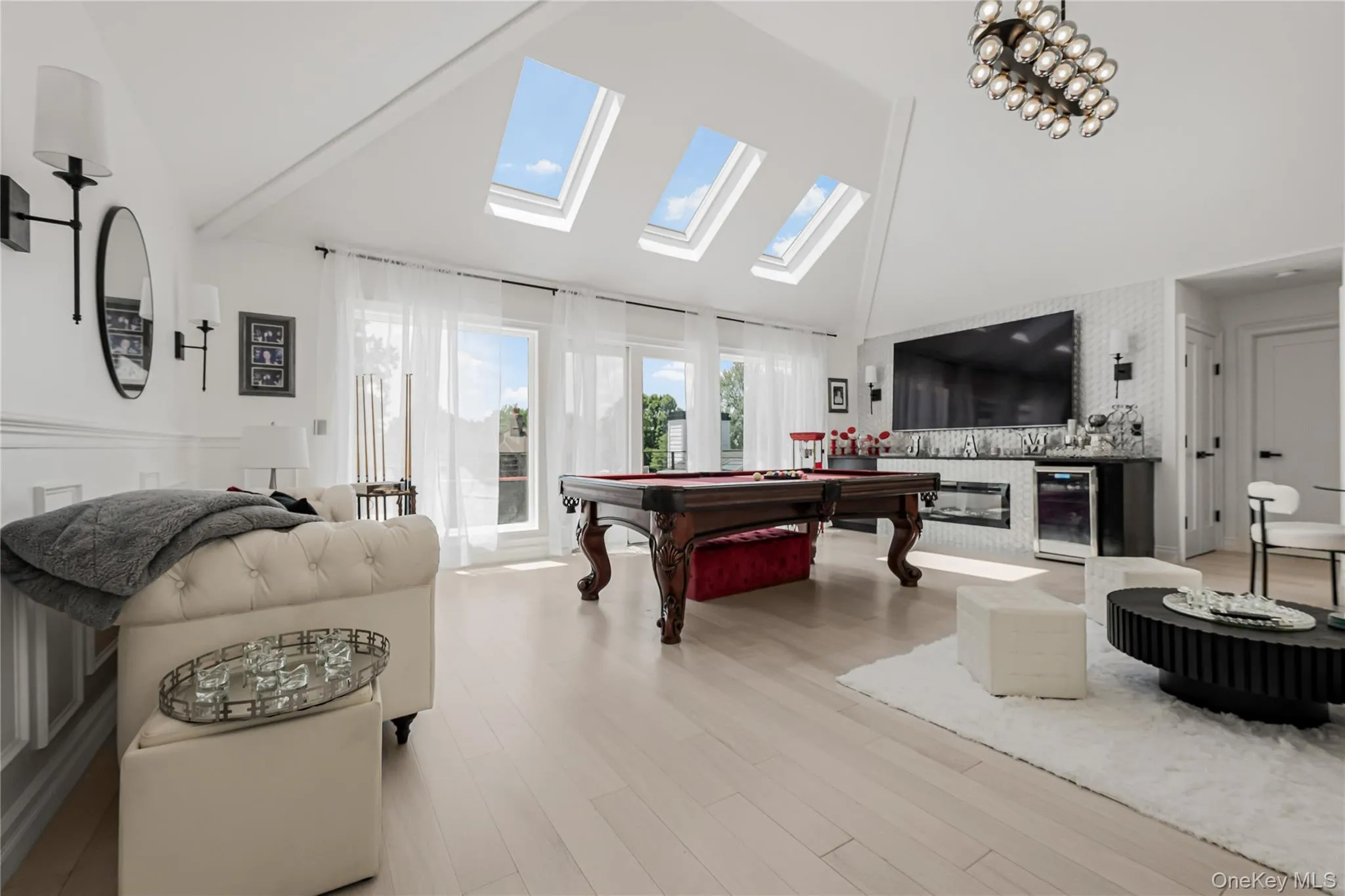 Game room featuring wine cooler, high vaulted ceiling, a skylight, light wood-style floors, and a glass covered fireplace Game room featuring wine cooler, high vaulted ceiling, a skylight, light wood-style floors, and a glass covered fireplace