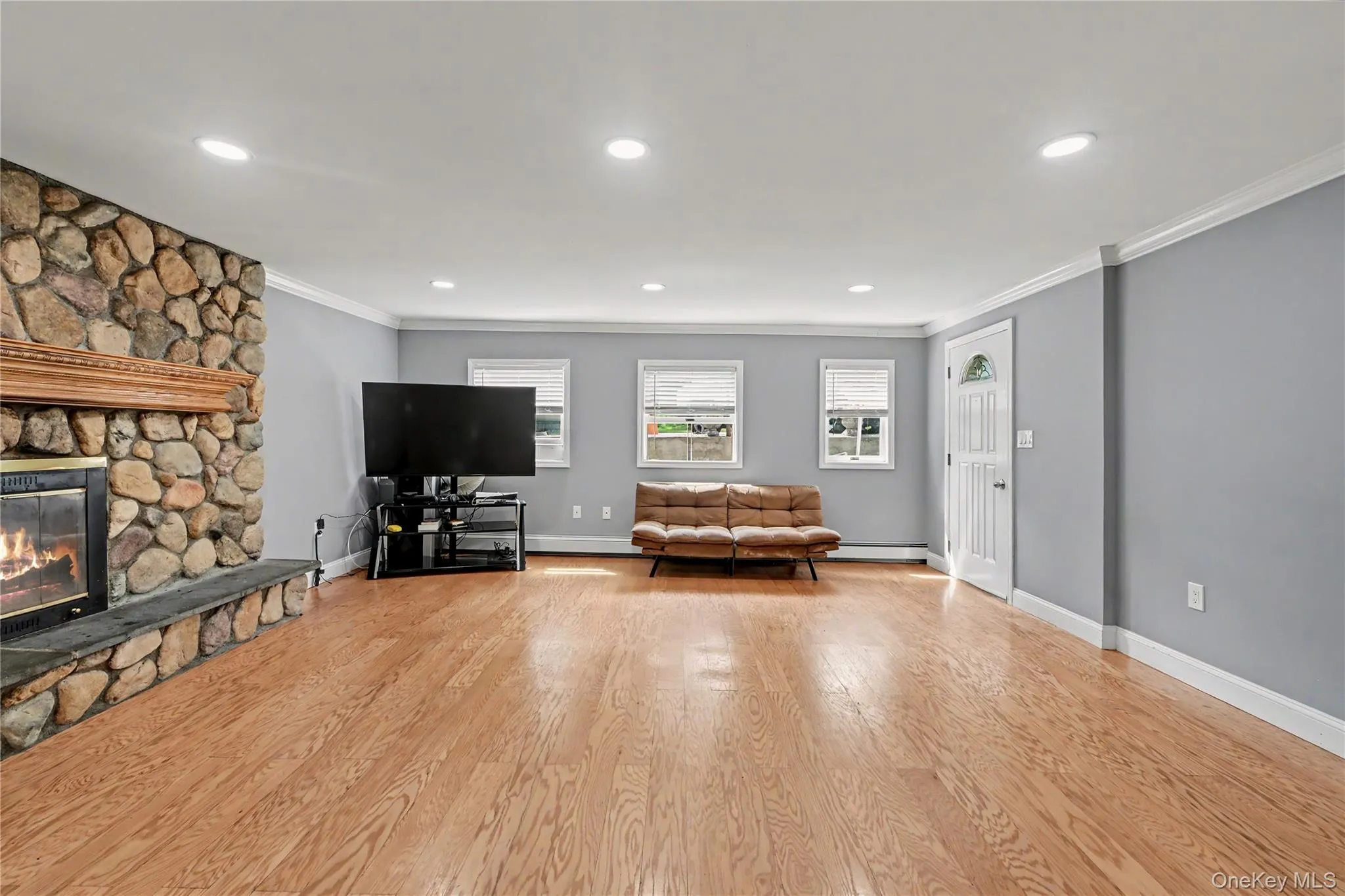 Spacious living area featuring polished wood-finish flooring, a stone-faced fireplace with a wood mantel, recessed lighting, and crown molding. Spacious living area featuring polished wood-finish flooring, a stone-faced fireplace with a wood mantel, recessed lighting, and crown molding.
