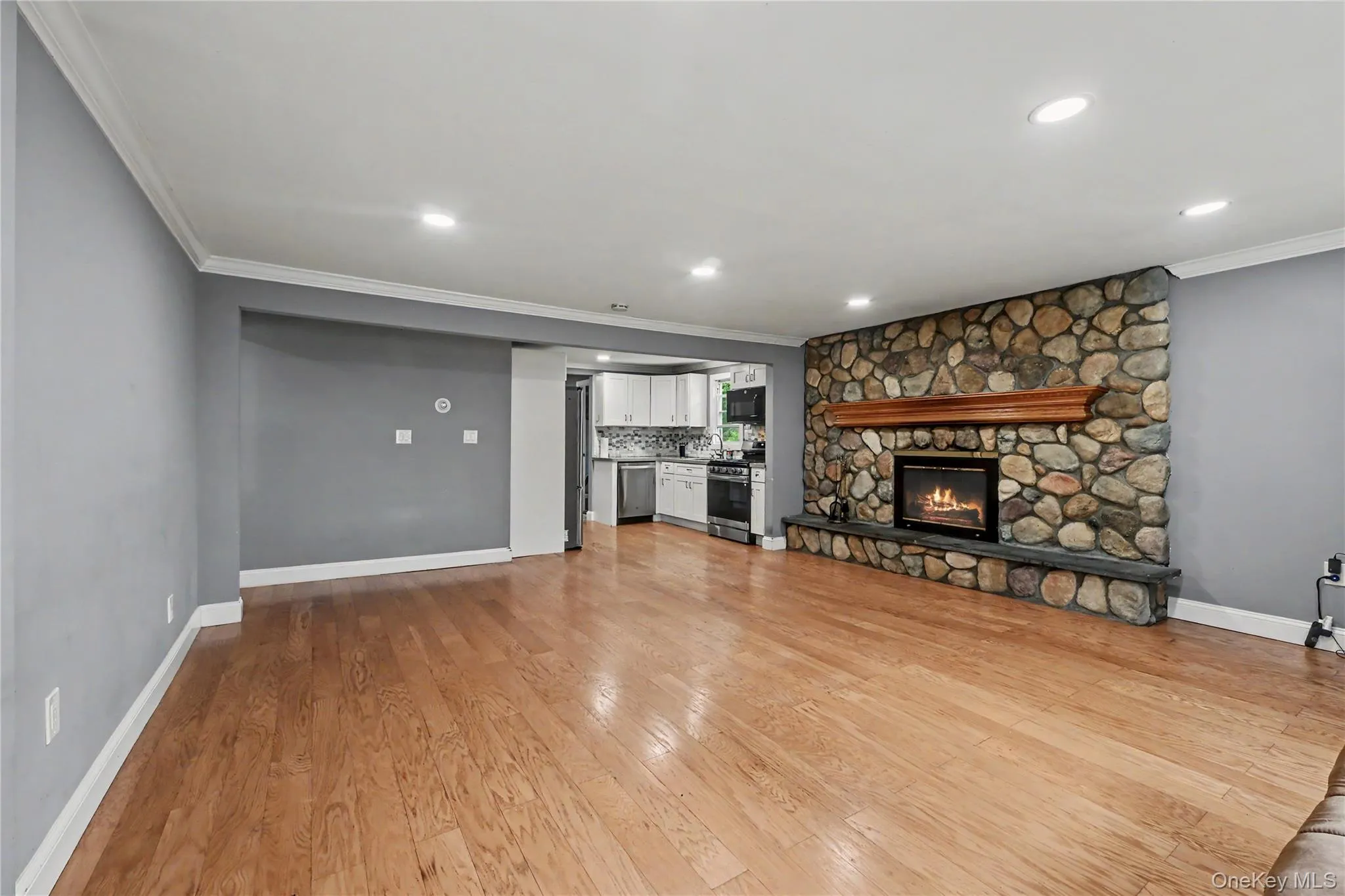 Spacious living area featuring hardwood flooring and a stone-clad fireplace with a wood mantle, View to kitchen. Spacious living area featuring hardwood flooring and a stone-clad fireplace with a wood mantle, View to kitchen.