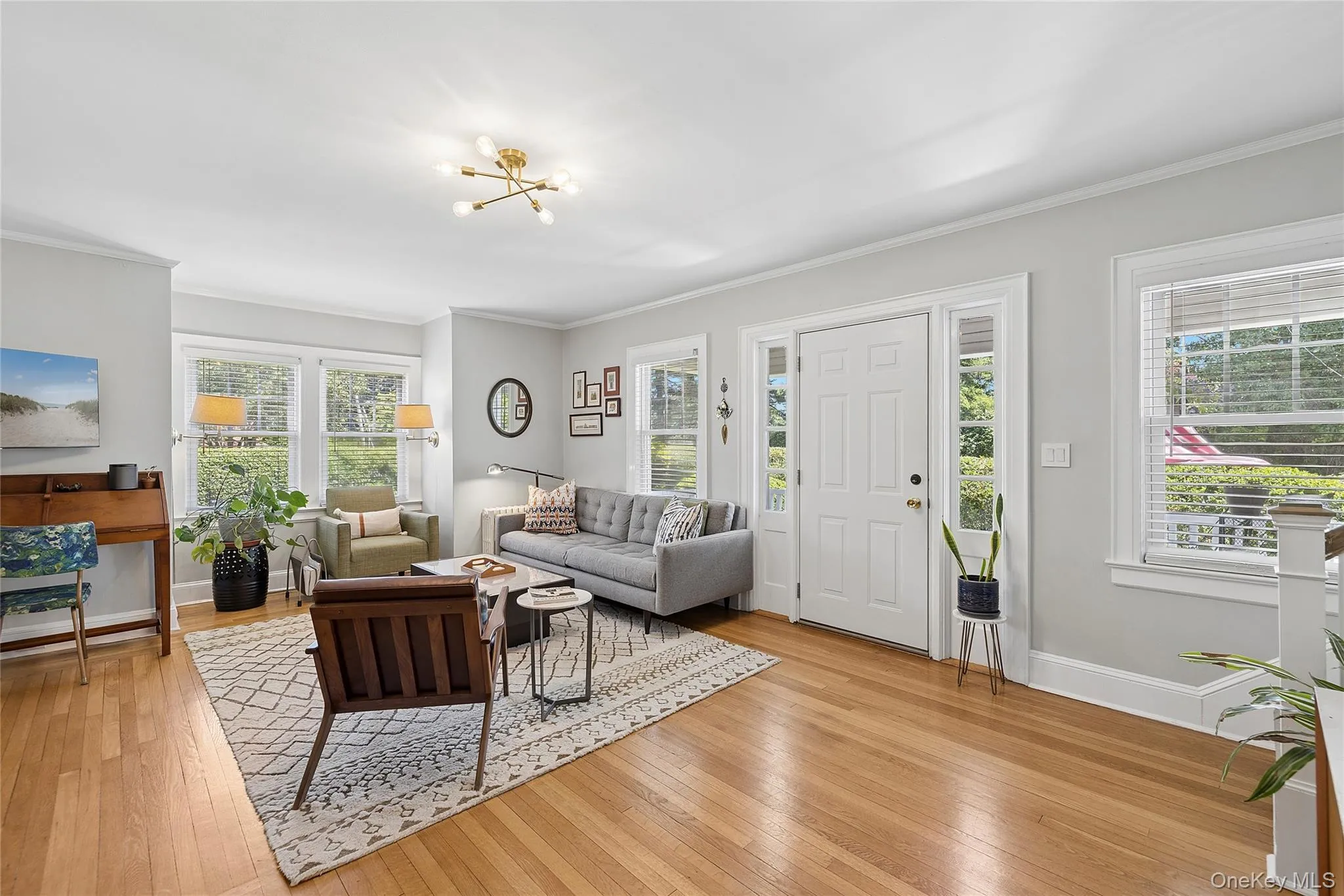 Light-filled living area featuring wood-finish flooring, a contemporary chandelier, multiple windows with natural light, and a white paneled entry door with sidelights Light-filled living area featuring wood-finish flooring, a contemporary chandelier, multiple windows with natural light, and a white paneled entry door with sidelights