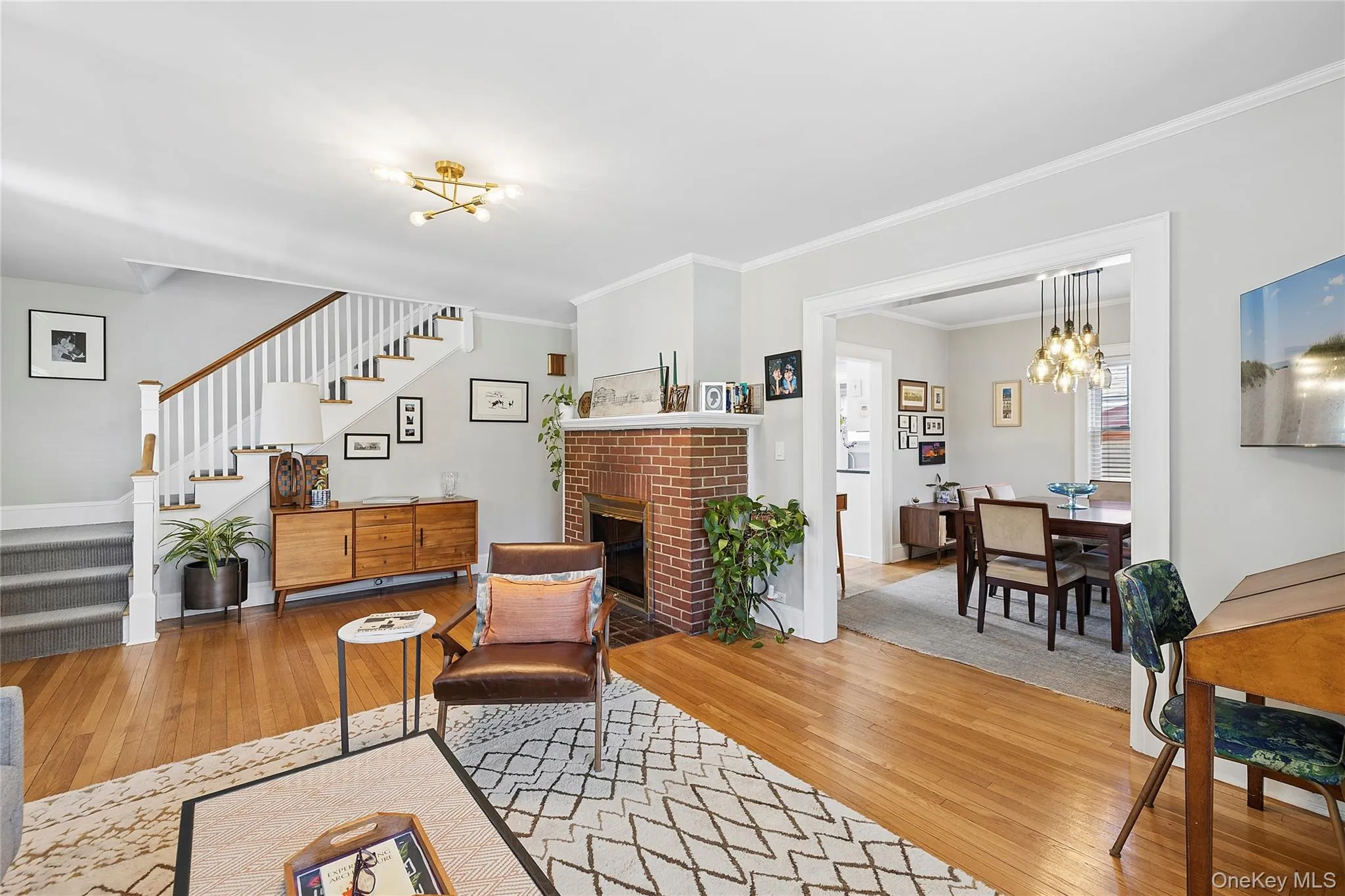 Spacious living area featuring warm wood-finish flooring, a white spindle staircase, and a brick fireplace Spacious living area featuring warm wood-finish flooring, a white spindle staircase, and a brick fireplace
