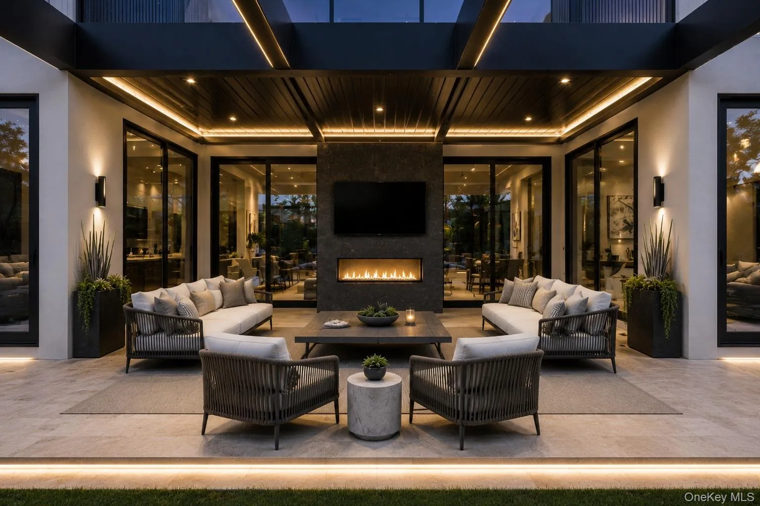 Expansive outdoor patio featuring a linear fireplace set into a dark stone accent wall, recessed ceiling lighting, and integrated strip lighting Expansive outdoor patio featuring a linear fireplace set into a dark stone accent wall, recessed ceiling lighting, and integrated strip lighting