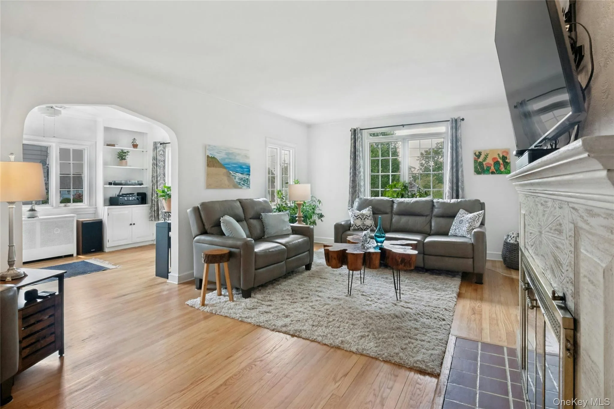 Living area with wood-finish flooring, a decorative fireplace with a tile hearth, and an arched entry to an alcove featuring built-in shelving and cabinetry Living area with wood-finish flooring, a decorative fireplace with a tile hearth, and an arched entry to an alcove featuring built-in shelving and cabinetry
