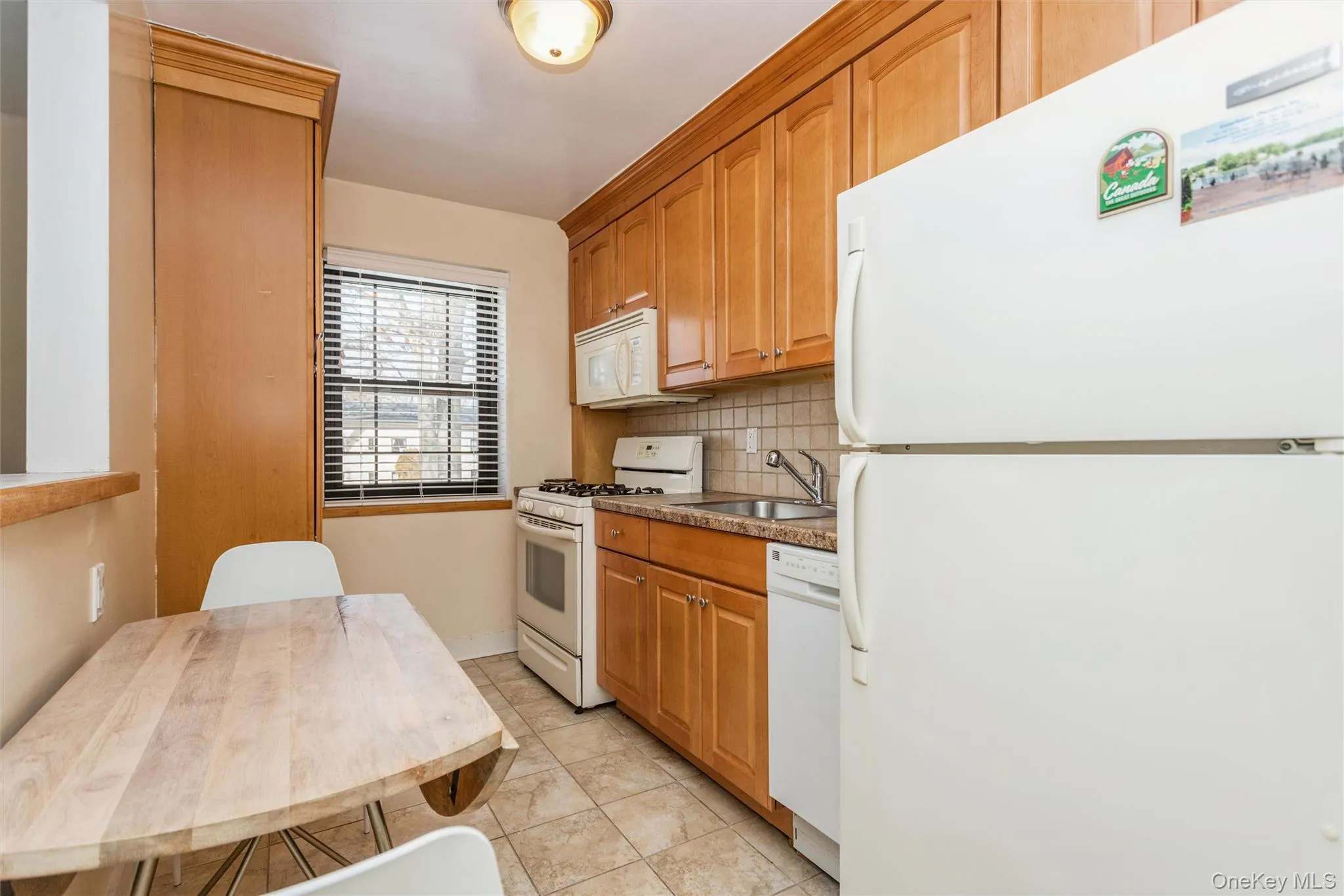 Kitchen featuring white appliances, backsplash, brown cabinetry, light tile patterned floors, and dark countertops Kitchen featuring white appliances, backsplash, brown cabinetry, light tile patterned floors, and dark countertops