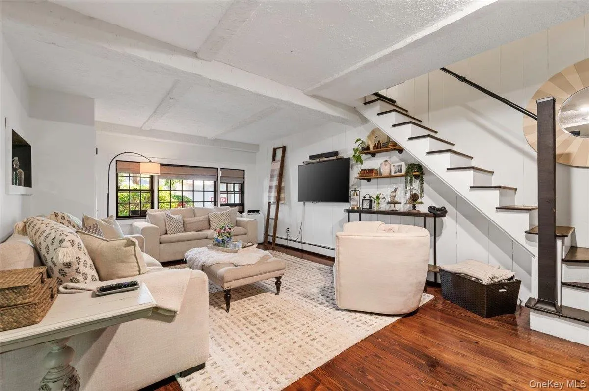 Living area featuring wood-finish flooring, exposed ceiling beams, and a modern staircase with dark treads and a black handrail Living area featuring wood-finish flooring, exposed ceiling beams, and a modern staircase with dark treads and a black handrail