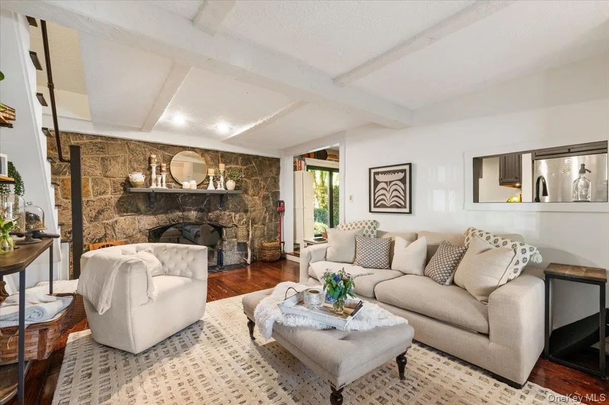 Spacious living area featuring a stone fireplace, wood-finish flooring, exposed ceiling beams, and a pass-through window Spacious living area featuring a stone fireplace, wood-finish flooring, exposed ceiling beams, and a pass-through window