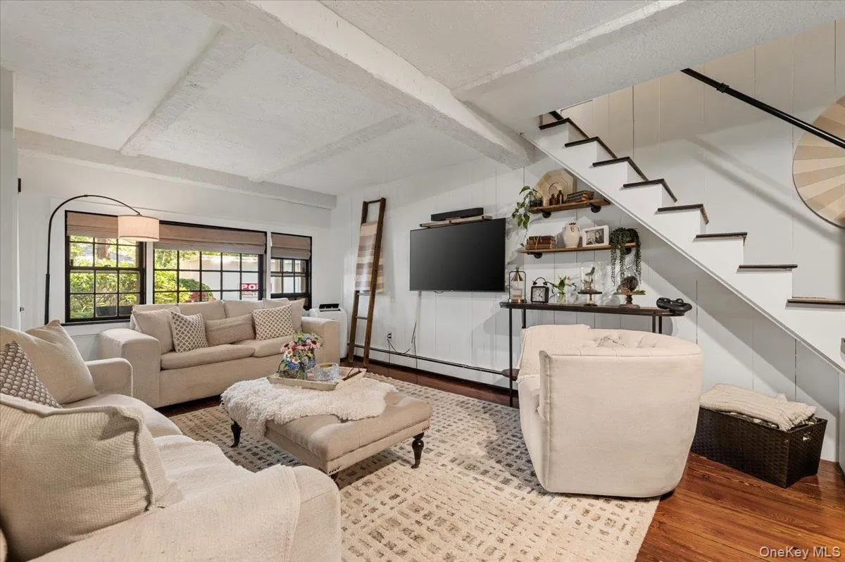 Open-concept living area featuring exposed ceiling beams, wood-finish flooring, and a staircase with painted risers Open-concept living area featuring exposed ceiling beams, wood-finish flooring, and a staircase with painted risers