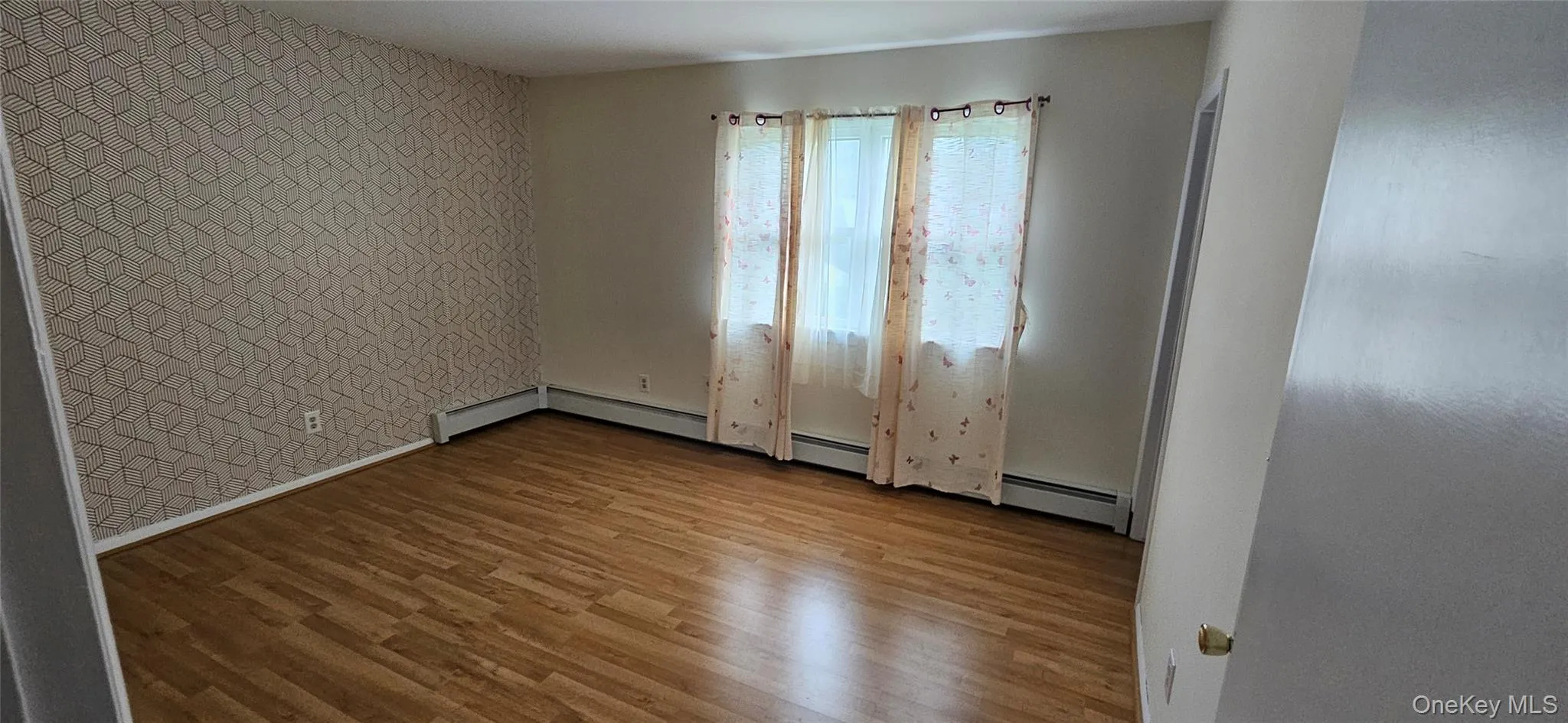 Spacious interior featuring wood-finish flooring, a geometric accent wall, and three windows Spacious interior featuring wood-finish flooring, a geometric accent wall, and three windows