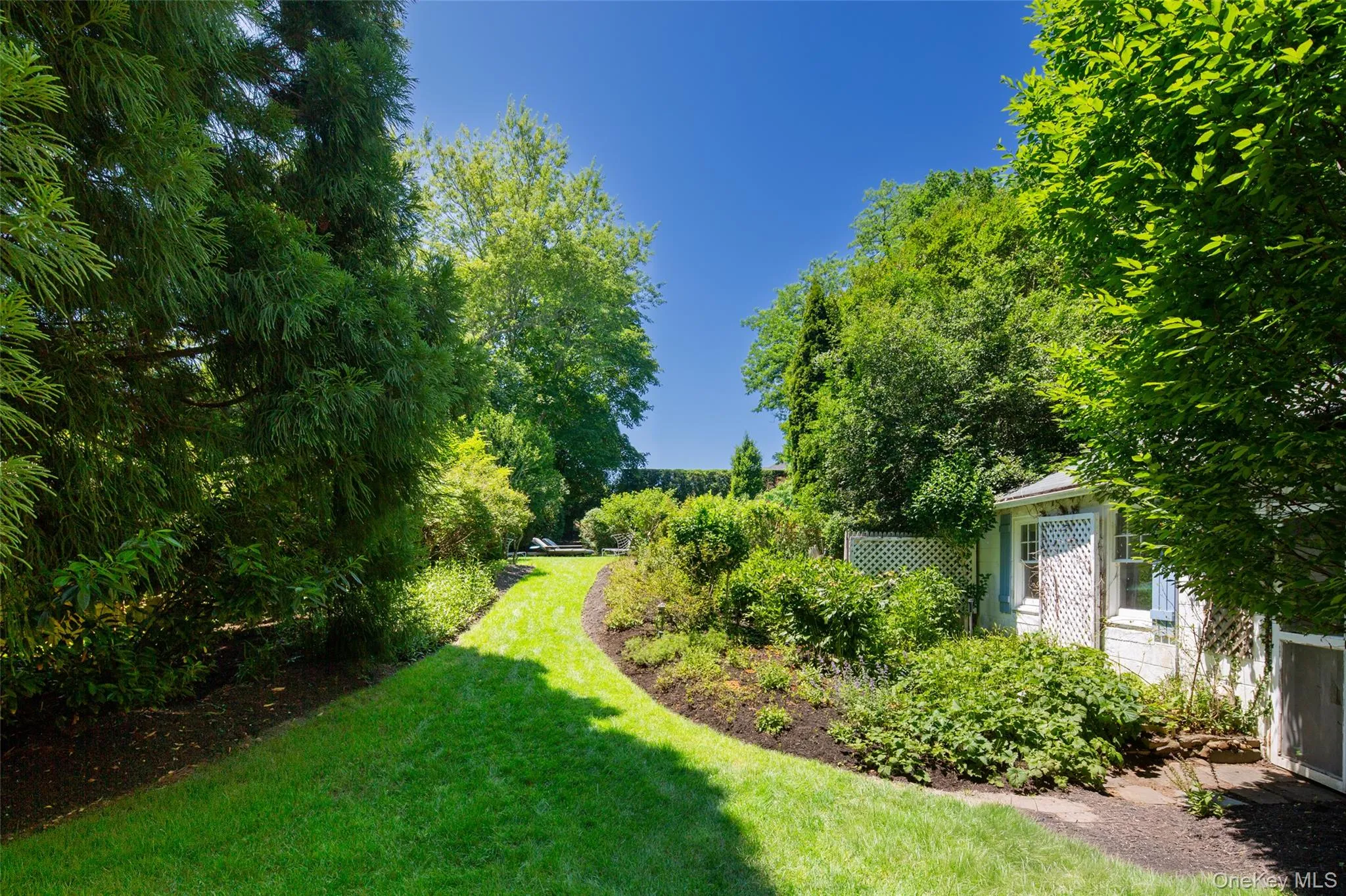 Landscaped grounds featuring a winding grassy path, mature trees, dense shrubbery, a white lattice fence, and a white shed with blue shutters Landscaped grounds featuring a winding grassy path, mature trees, dense shrubbery, a white lattice fence, and a white shed with blue shutters