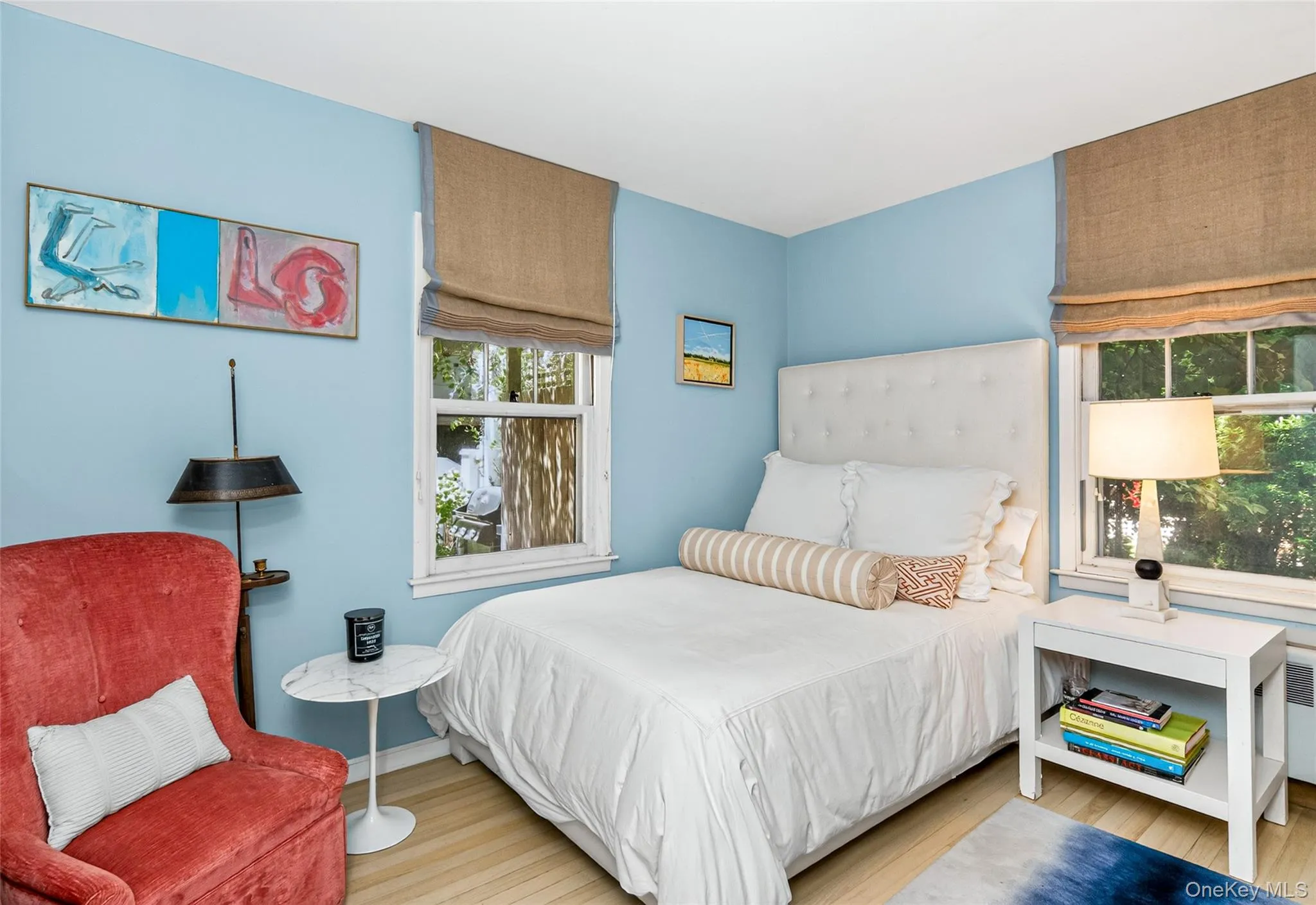 First Floor Bedroom featuring light wood-finish flooring, two large windows with natural fiber window treatments, a tufted white headboard, and light blue wall paint First Floor Bedroom featuring light wood-finish flooring, two large windows with natural fiber window treatments, a tufted white headboard, and light blue wall paint