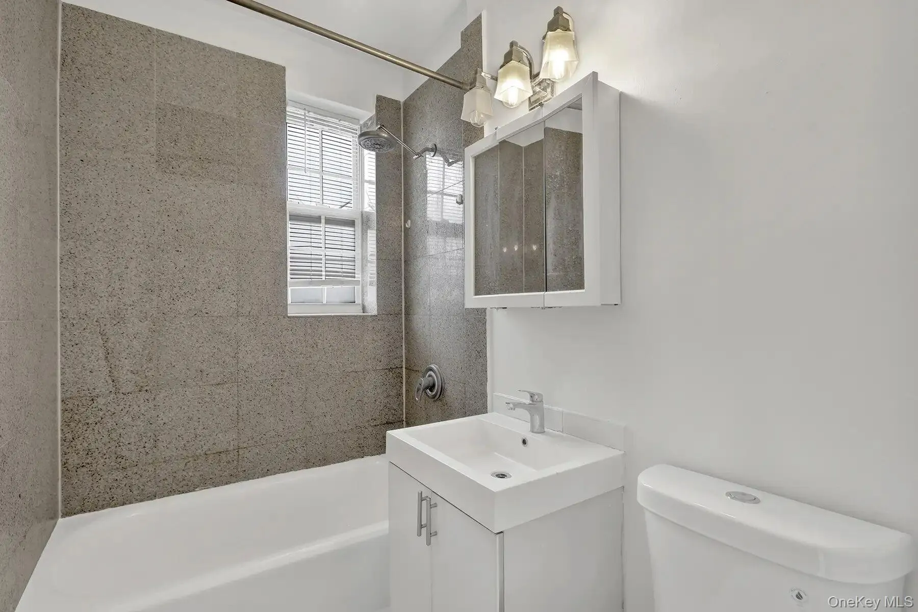 Bathroom featuring a white bathtub, gray tiled shower surround, and a window with blinds Bathroom featuring a white bathtub, gray tiled shower surround, and a window with blinds