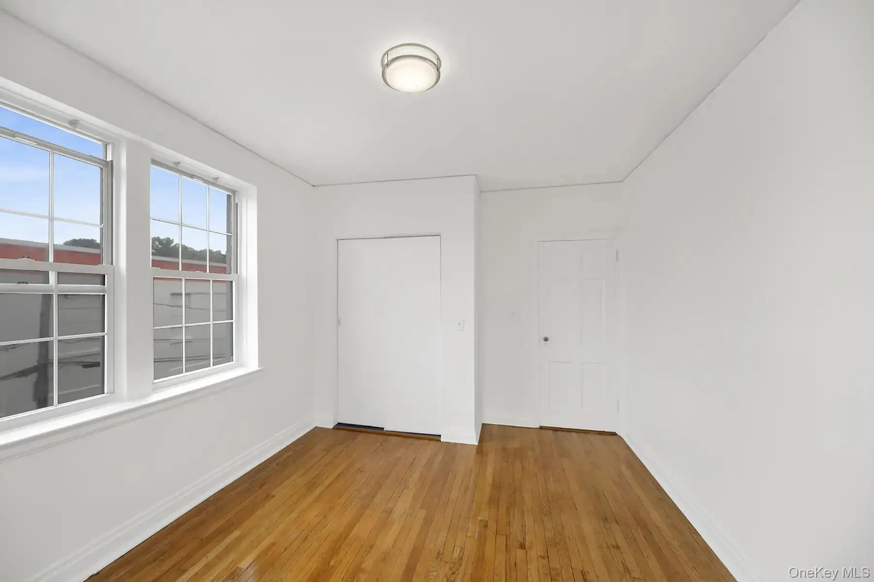 Room featuring wood-finish flooring, white baseboards, two multi-pane windows, a flush-mount ceiling light fixture, and two interior doors Room featuring wood-finish flooring, white baseboards, two multi-pane windows, a flush-mount ceiling light fixture, and two interior doors