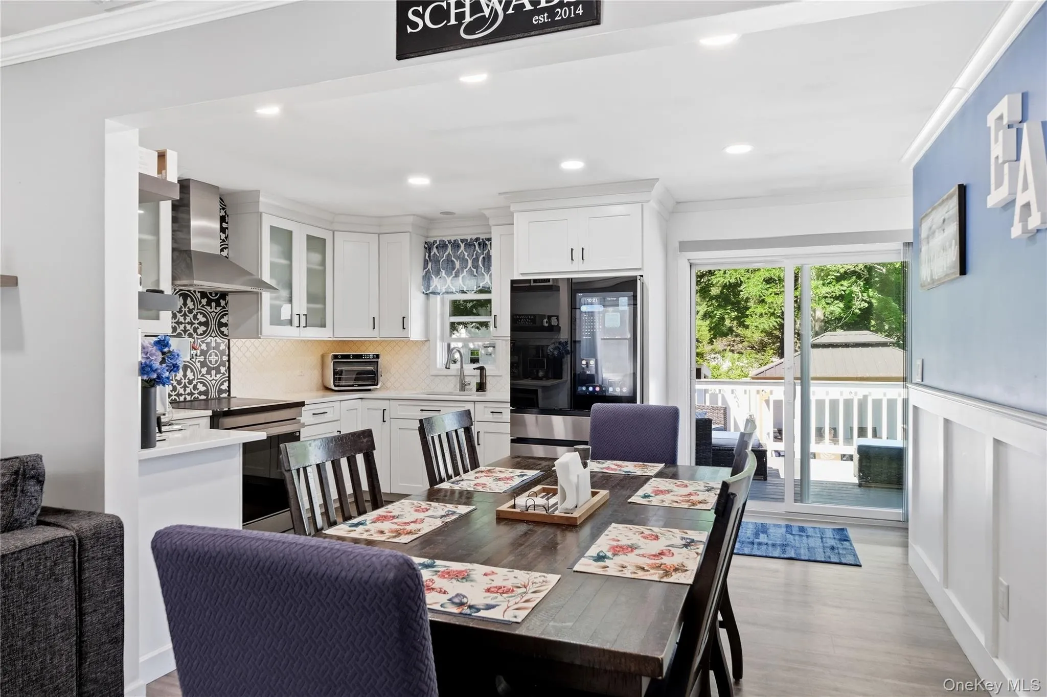 Open-concept living space featuring wood-finish flooring, a contemporary kitchen with white cabinetry and stainless steel appliances, and a dining area with direct access to an outdoor deck Open-concept living space featuring wood-finish flooring, a contemporary kitchen with white cabinetry and stainless steel appliances, and a dining area with direct access to an outdoor deck