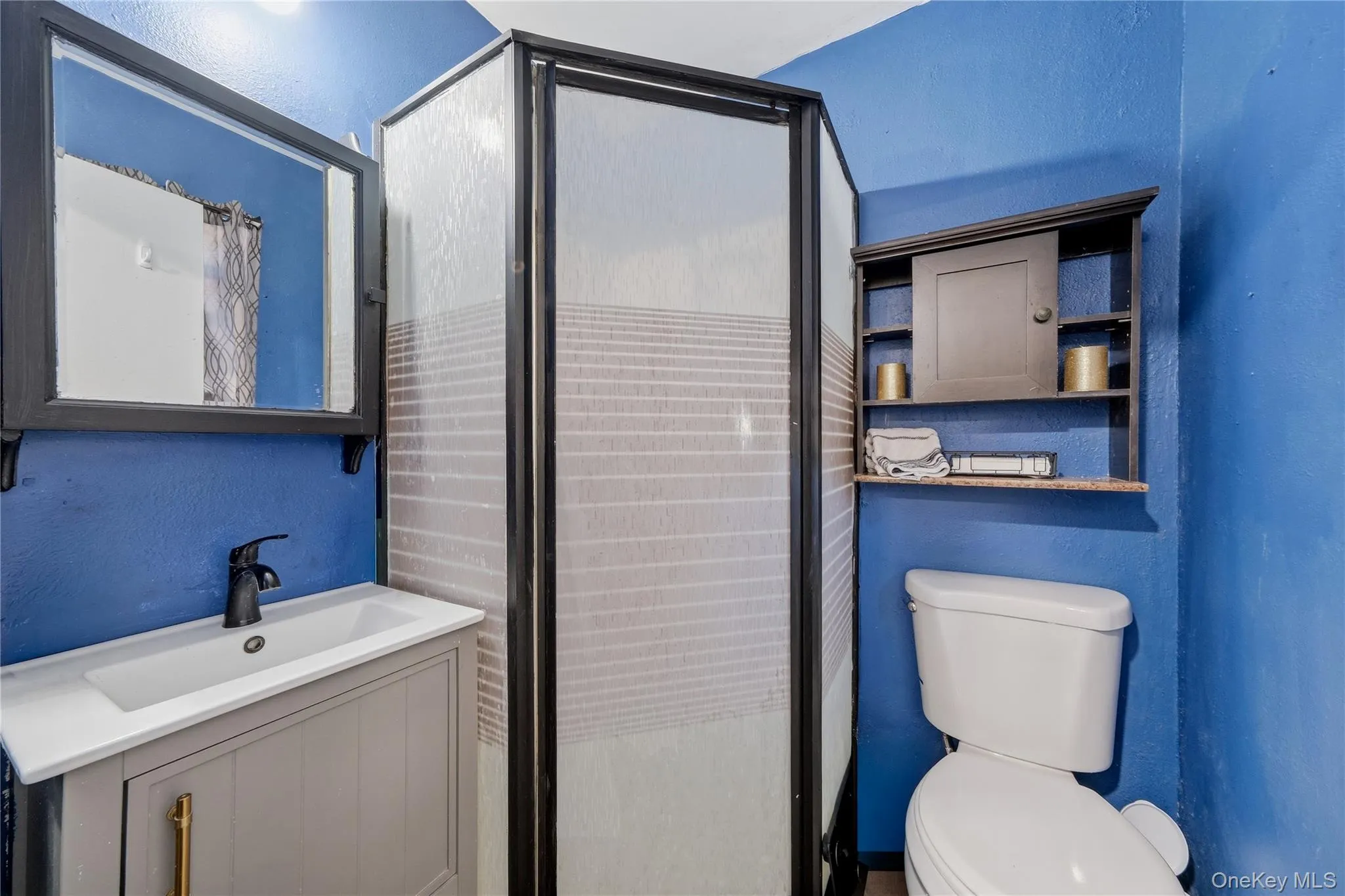 Compact bathroom featuring a corner shower enclosure with patterned glass, a vanity with a white rectangular sink and black faucet, a framed mirror, a wall-mounted storage cabinet, and a toilet Compact bathroom featuring a corner shower enclosure with patterned glass, a vanity with a white rectangular sink and black faucet, a framed mirror, a wall-mounted storage cabinet, and a toilet