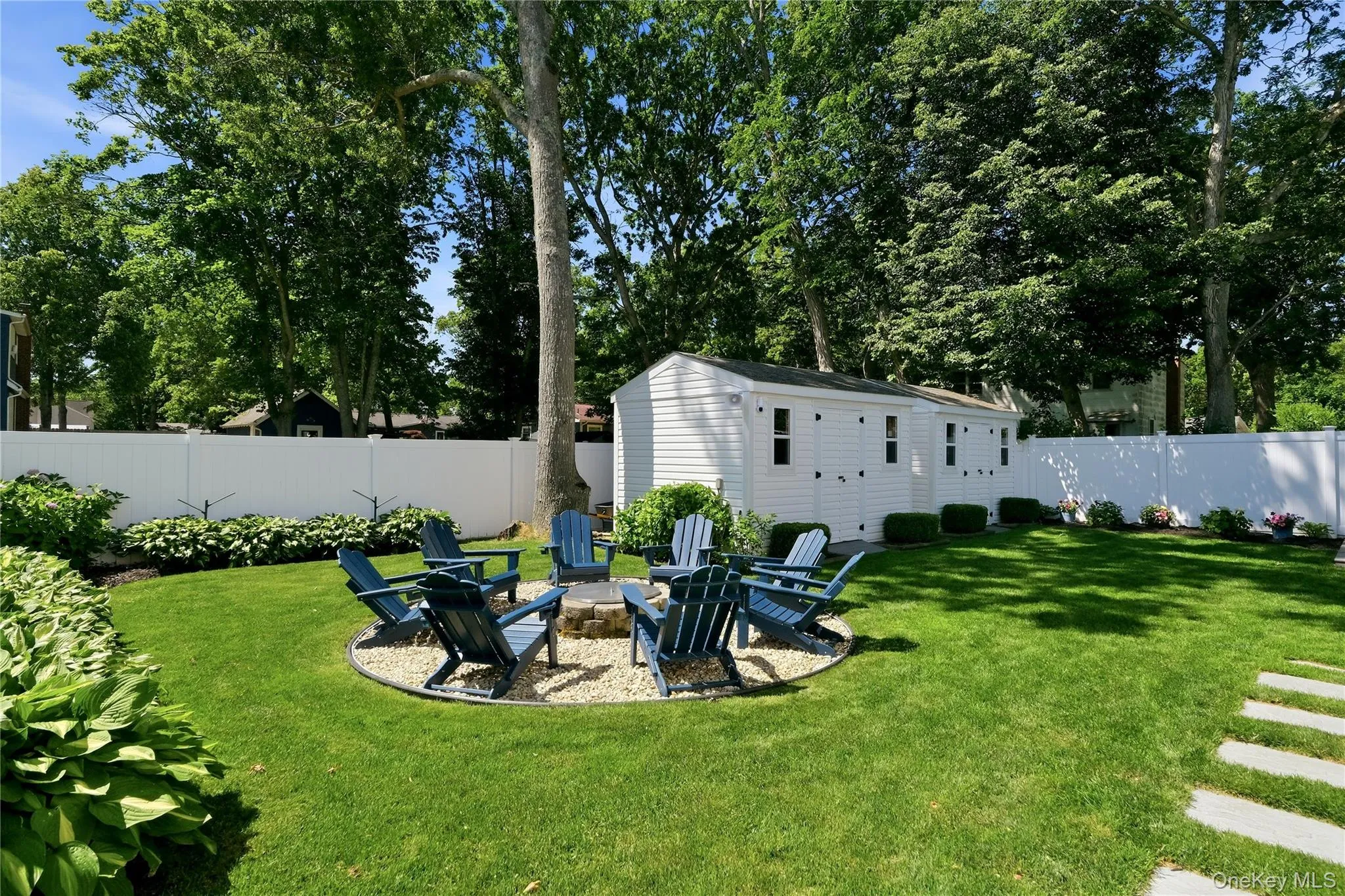 Landscaped backyard featuring a pea gravel fire pit area, expansive lawn, mature tree, white vinyl fencing, and a detached white storage shed Landscaped backyard featuring a pea gravel fire pit area, expansive lawn, mature tree, white vinyl fencing, and a detached white storage shed