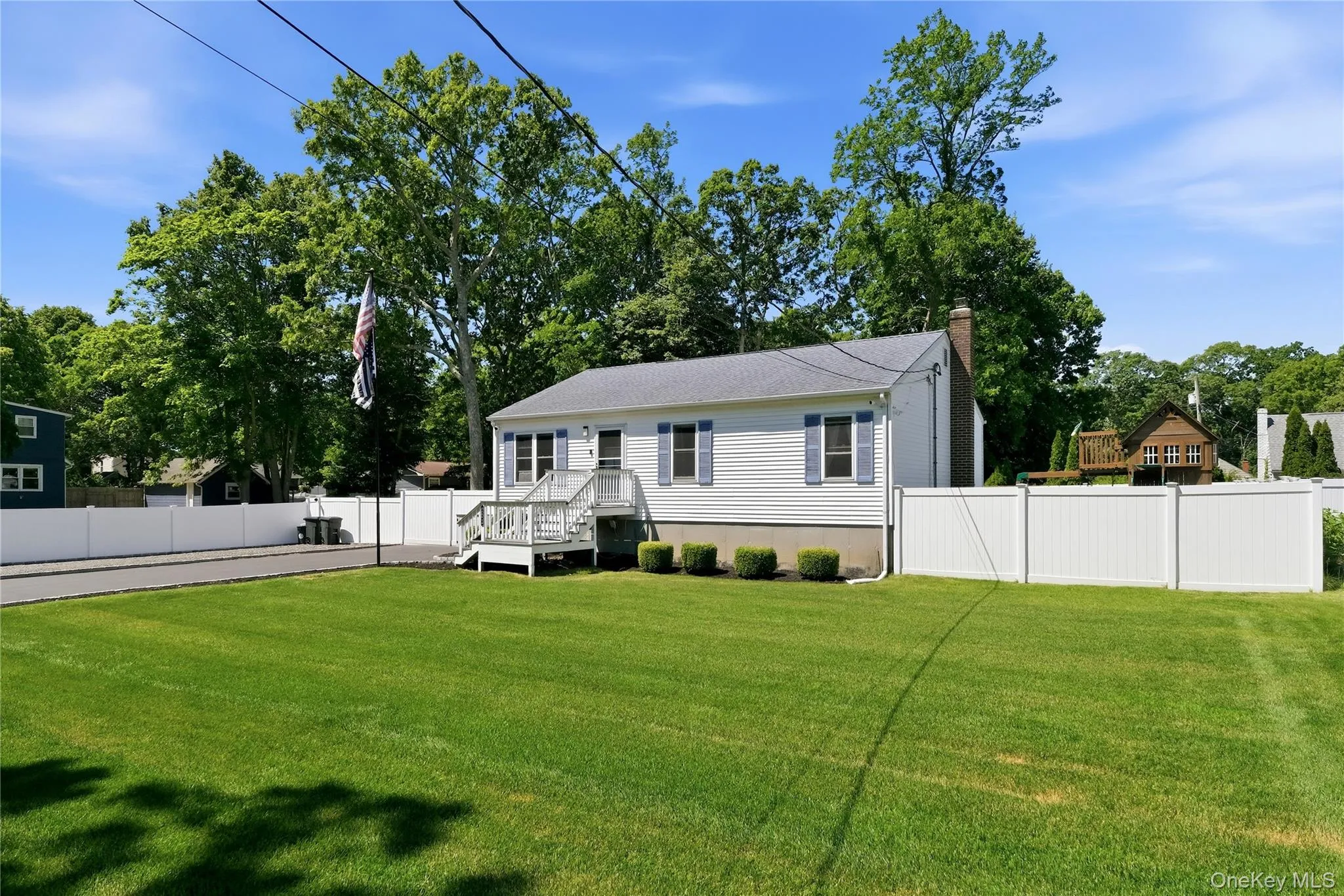 White siding residence with a dark shingle roof, featuring blue shutters and a prominent front deck with steps White siding residence with a dark shingle roof, featuring blue shutters and a prominent front deck with steps