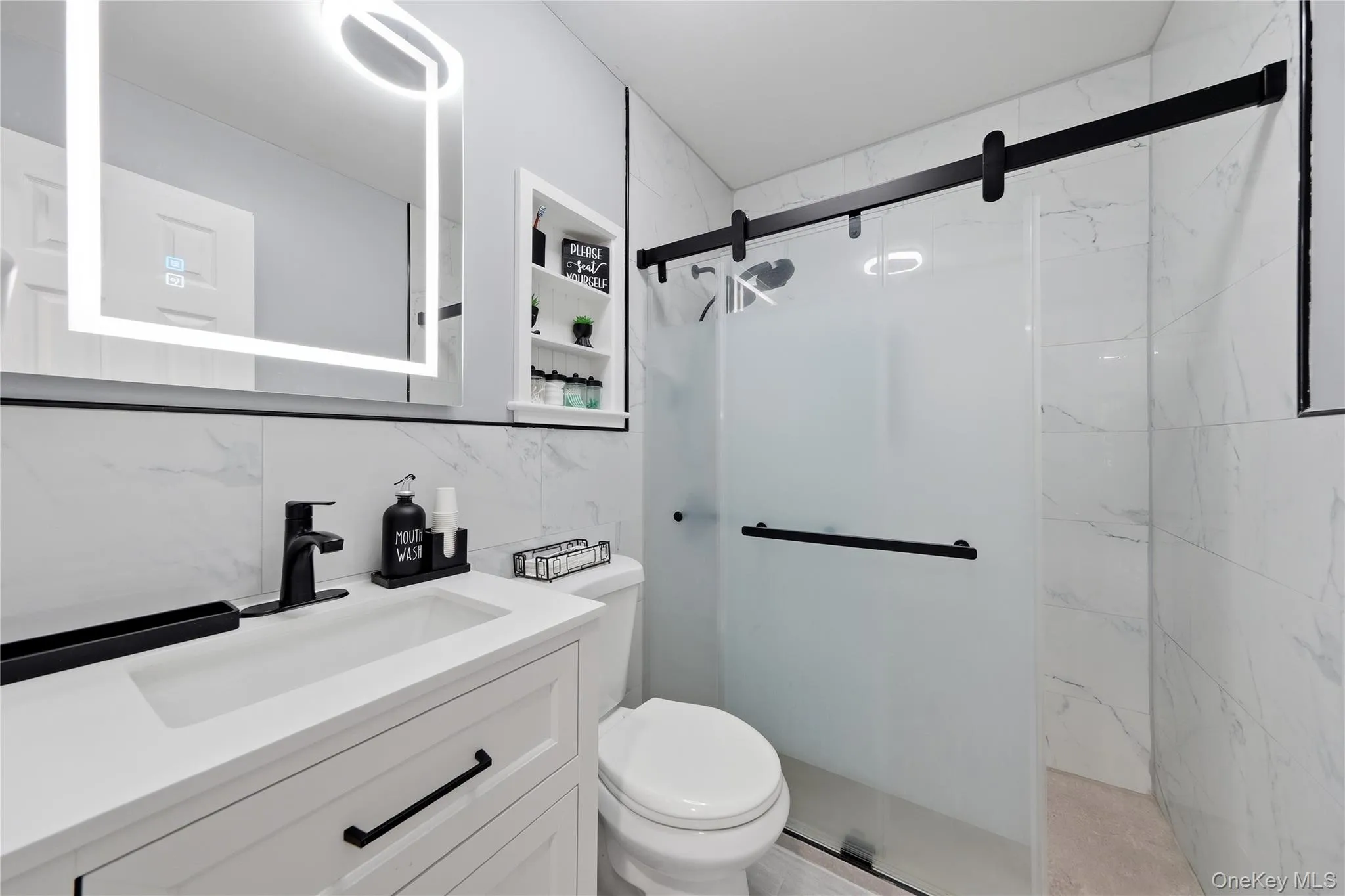 Modern bathroom featuring a white vanity with integrated sink, black hardware, an illuminated mirror, and a frosted glass shower enclosure with a barn door style track Modern bathroom featuring a white vanity with integrated sink, black hardware, an illuminated mirror, and a frosted glass shower enclosure with a barn door style track